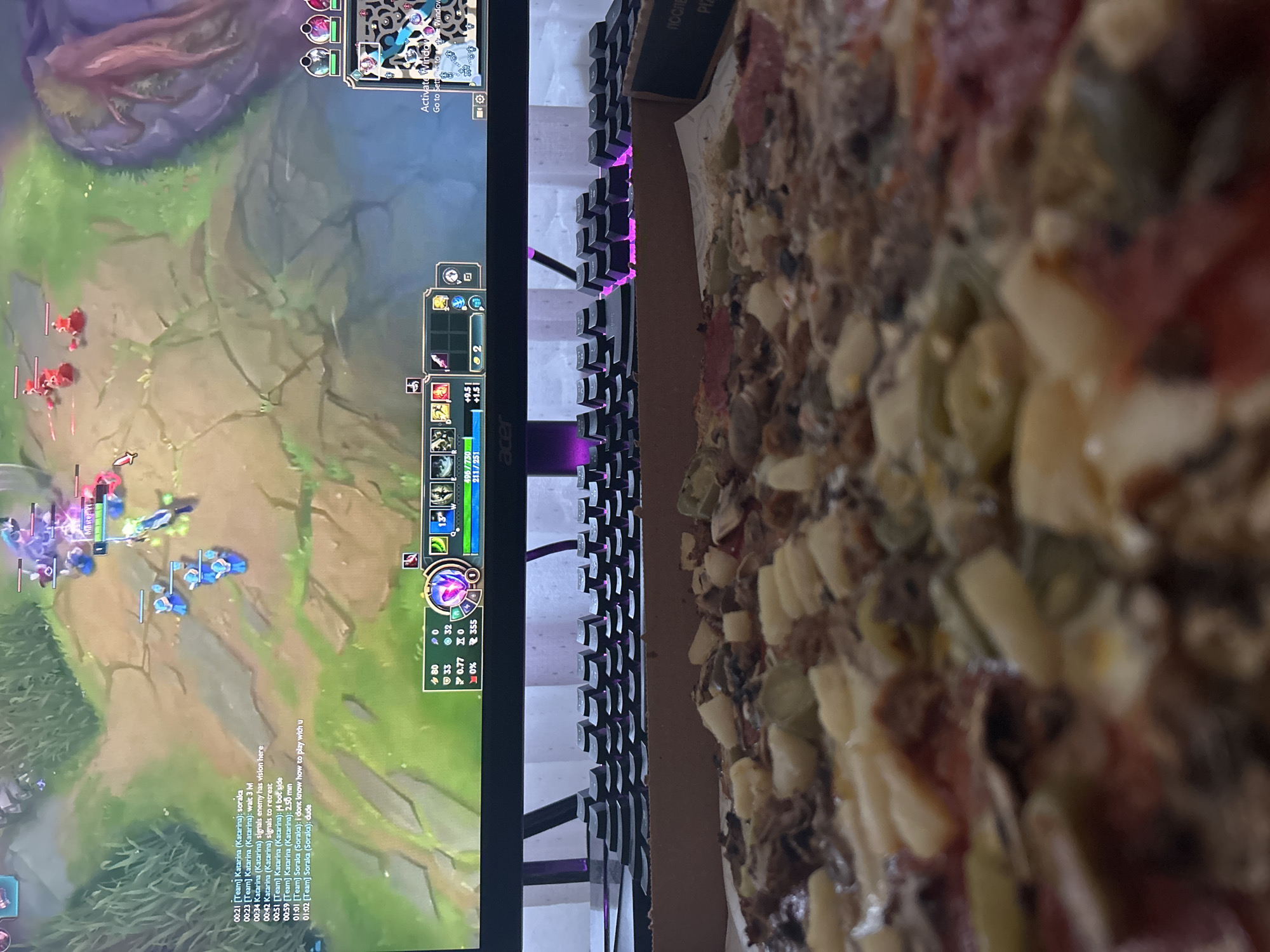 The image, rotated 90 degrees clockwise, presents an indoor scene with a computer setup and food. On the left, a computer monitor displays a video game, identifiable as League of Legends by its user interface elements and gameplay. The screen shows a jungle area with champions and minions, along with a minimap, character statistics (e.g., "496/730," "211/251," "33" kills, "32" assists), and a chat window. The chat log includes messages from players named "Katarina" and "Soraka," discussing in-game strategy and expressing frustration, such as "soraka," "wait 3 IM," "signals enemy has vision here," "signals to retreat," "14 bot side," "2.50 min," "i dont know how to play with u," and "dude." The monitor features an "acer" logo and a "Activate Windows" watermark.
Positioned between the monitor and the food is a black mechanical keyboard with purple backlighting.
To the right, a close-up view of a pizza in a cardboard box is visible. The pizza is loaded with various toppings, including distinct pineapple chunks, slices of salami or pepperoni, and other finely diced ingredients. Text on the pizza box flap reads "ПОСЛЕ" (Cyrillic for 'After' or 'Later') and partially "Pla". The overall scene depicts a person engaged in gaming, likely consuming pizza, in a casual setting.