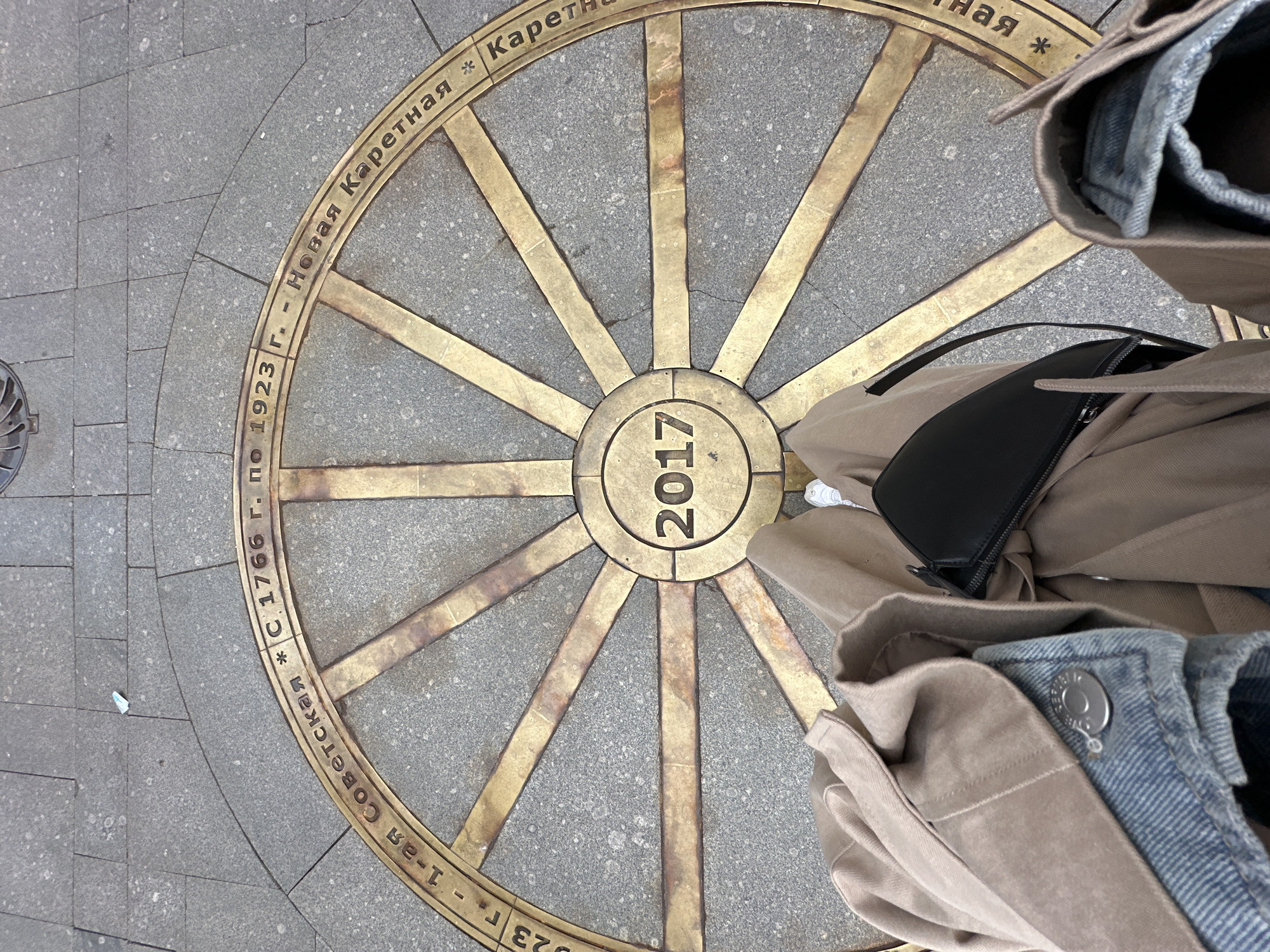 A high-angle shot depicts a circular, golden marker embedded in the pavement, bearing the year "2017" in its center, with radial lines extending outwards like spokes of a wheel. The text "Новая Каретная" is visible, along with "С 1766 г. по 1923 г. - 1-ая Советская" around the perimeter. A person's legs and a black purse are visible on the right side of the frame. The location is likely in Saint Petersburg, Russia, identified by the Cyrillic script on the marker. The marker seems to commemorate the history or renaming of a street, with dates indicating historical periods.