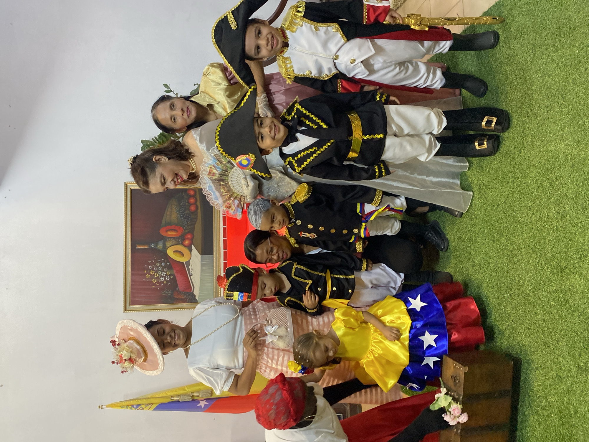 This indoor scene, located in Tinaquillo, Venezuela, features ten individuals—five adults and five children—posed for a photograph on a green artificial turf floor against a plain white wall.
On the left, a Venezuelan flag stands erect. A gold-framed still-life painting, depicting fruits and bottles, hangs centrally on the wall.
Five adult women are present. One woman, positioned left-center, wears a white blouse, a pink striped skirt, a long pearl necklace, and a large, decorated pink hat. She holds a small white handbag. Another woman, central, wears a light-colored historical gown and holds an open fan. A third woman, right-center, wears a gold blouse. A fourth woman, at the far left, wears a red headscarf and white top, bending towards a small wooden chest adorned with pink flowers. A fifth woman, on the right, wears glasses and a dark top, kneeling behind two children.
Five children are also present, dressed in historical costumes. One young girl, at the front-left, wears a yellow dress with a blue skirt featuring white stars and a red underskirt. Four young boys are dressed in military-style uniforms from the independence era, characterized by bicorne hats, gold braiding, white trousers, and dark boots. One boy on the far right holds a gold-colored toy sword. Most individuals are looking towards the camera; several adults and children are smiling.