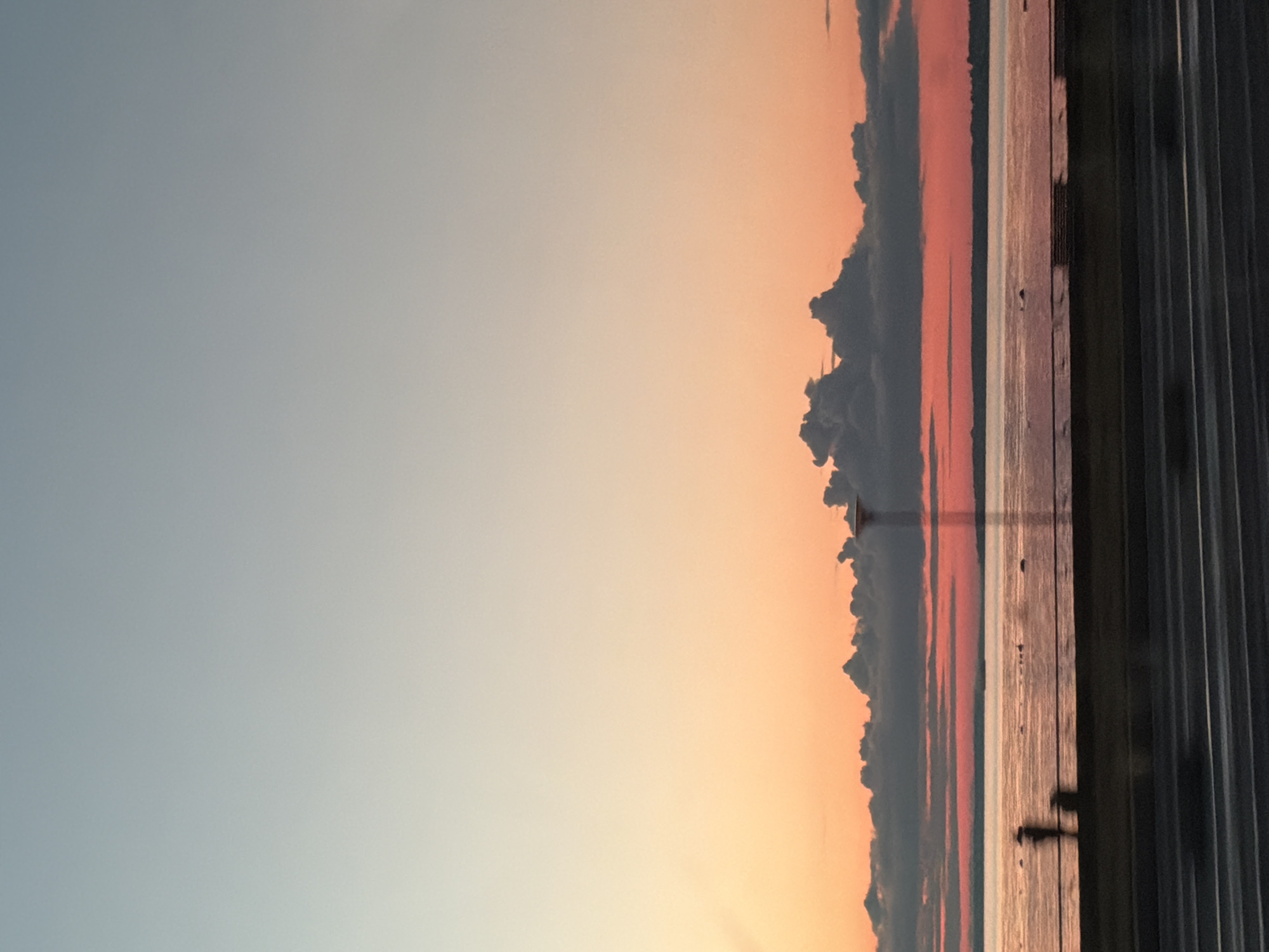 The image captures a sunset over the Baltic Sea in Tallinn, Estonia. The sky transitions from a pale blue at the top to a soft orange and pink hue near the horizon. Dark, dramatic clouds are visible against the colorful sky. The sea reflects the warm tones of the sunset, creating a shimmering surface. In the foreground, a silhouetted figure stands near the water's edge, with another indistinct figure nearby. A lamppost stands to the right of the silhouettes. A road with white markings runs along the bottom of the image, suggesting the scene is viewed from a roadside or a vehicle.