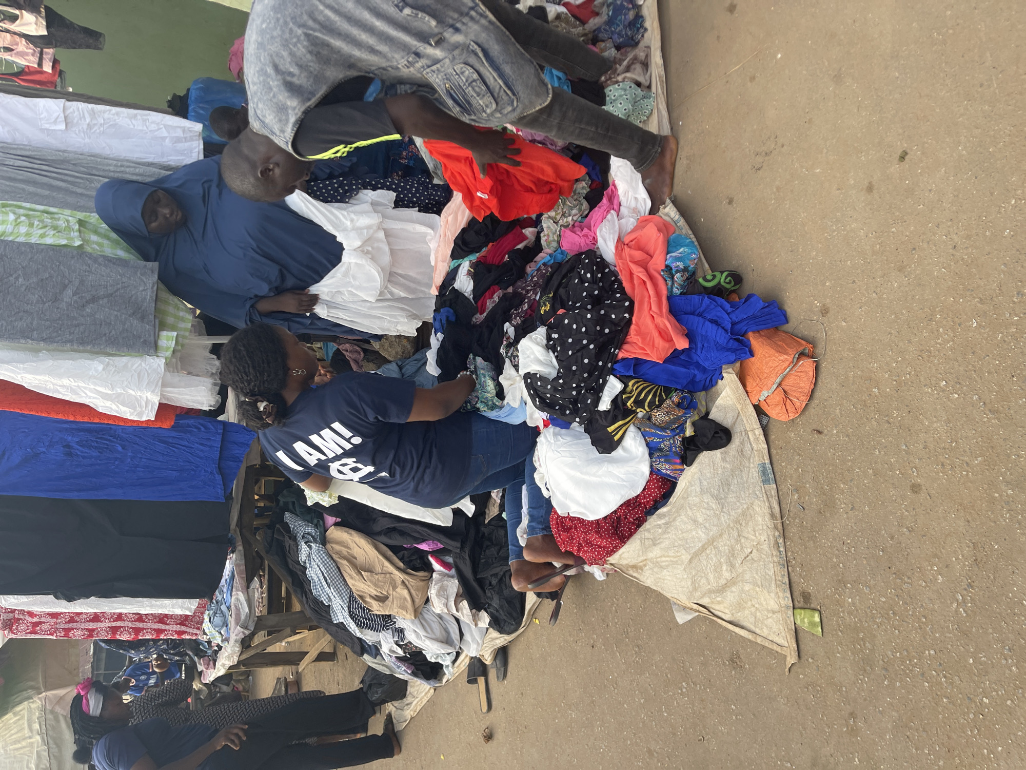 The image shows an outdoor scene in what appears to be Akwanga, Nigeria. Multiple people are gathered around a large pile of clothes laid out on a tarpaulin on the ground. Several individuals are sifting through the clothing, presumably browsing or selecting items. The clothes appear to be a mix of colors, patterns, and materials. Two of the people at the stand seem to be selling clothing. One is a woman wearing a blue hijab, another is standing barefoot and sorting clothes. The ground is paved, suggesting an urban or market setting. Additional garments, neatly stacked, are visible on a raised platform or table-like structure to the side, likely part of the display or for storage.
