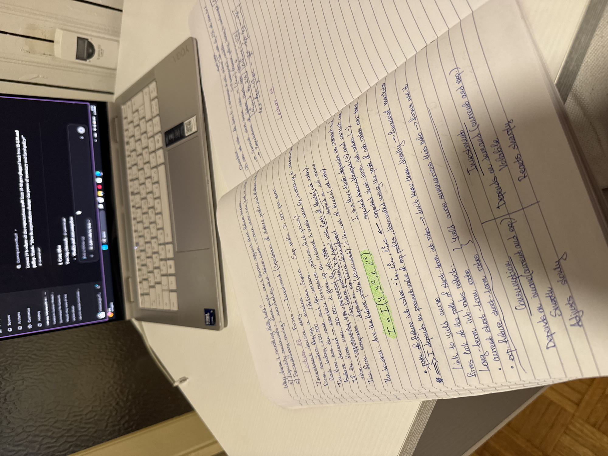 Here's a forensic description of the image:
**Setting:** An indoor setting, likely a home or office desk area.
**Objects:**
* A silver Lenovo Yoga laptop is open and powered on. The screen displays a conversation with an AI or chatbot program.
* A spiral-bound notebook is open, filled with handwritten notes in blue ink. The notes appear to be economics or finance-related, containing formulas and diagrams.
* A small, white electrical outlet or switch is mounted on the wall in the background.
* A wood floor is in the very foreground of the scene.
**Subjects:**
* The notes on the notebook appear to be handwritten, indicating a human presence.
* The AI interface on the laptop indicates a user interacting with the computer.
**Actions/Interactions:**
* Someone is taking notes, seemingly while also referencing information on the laptop screen.
* The AI interface displays a text-based conversation.
**Environment:**
* The lighting in the scene is soft, likely from natural or ambient light sources.
* The environment seems clean and organized.
**Inference:**
Based on the contents of the handwritten notes, the laptop screen, and the overall setting, the scene likely depicts a student studying economics or finance using their laptop and notebook.
**Location:** There is no indication that the scene is in Milan, Italy.