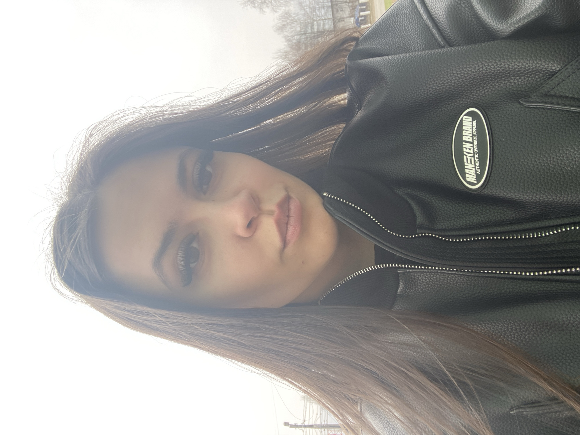 The image is a close-up selfie of a young woman with long, brown hair wearing a black leather jacket. The jacket has a patch that reads "MANTEEN BRAND AUTHENTIC OVERSIZE APPAREL". The background is blurry and bright, suggesting an outdoor location. Based on visual assessment, the image lacks definitive elements that would confirm its location within Chaikovsky, Russia.