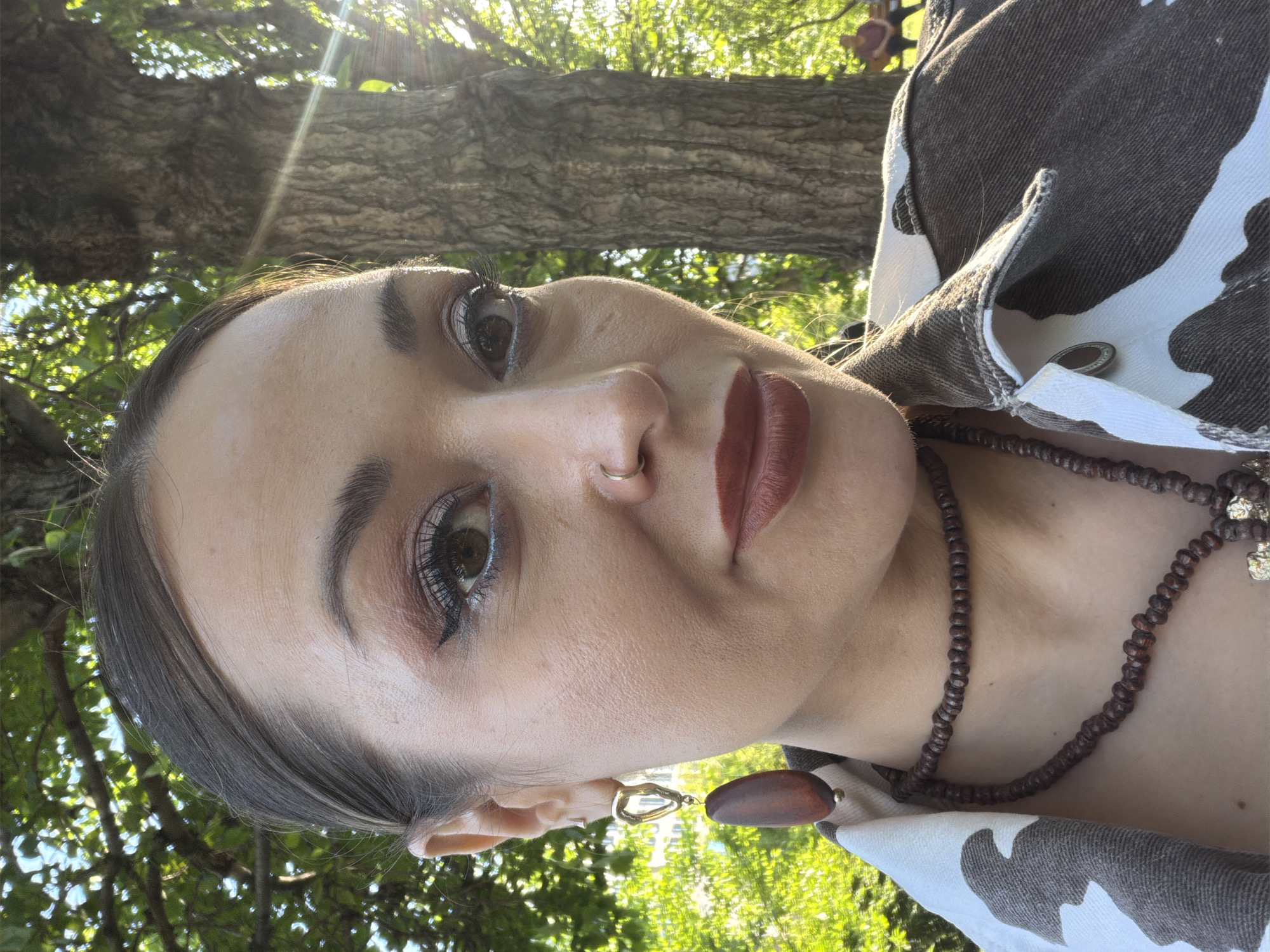 A close-up, rotated portrait features a person with light skin, brown eyes, and brown hair pulled back. They wear a nose ring, brown lipstick, and eye makeup. A brown beaded necklace and earrings with geometric gold shapes and brown, teardrop pendants are visible. The person is wearing a jacket with a black and white cow pattern. The background shows green leaves and a tree trunk. The location cannot be determined to be Krasnoyarsk, Russia, from the image.