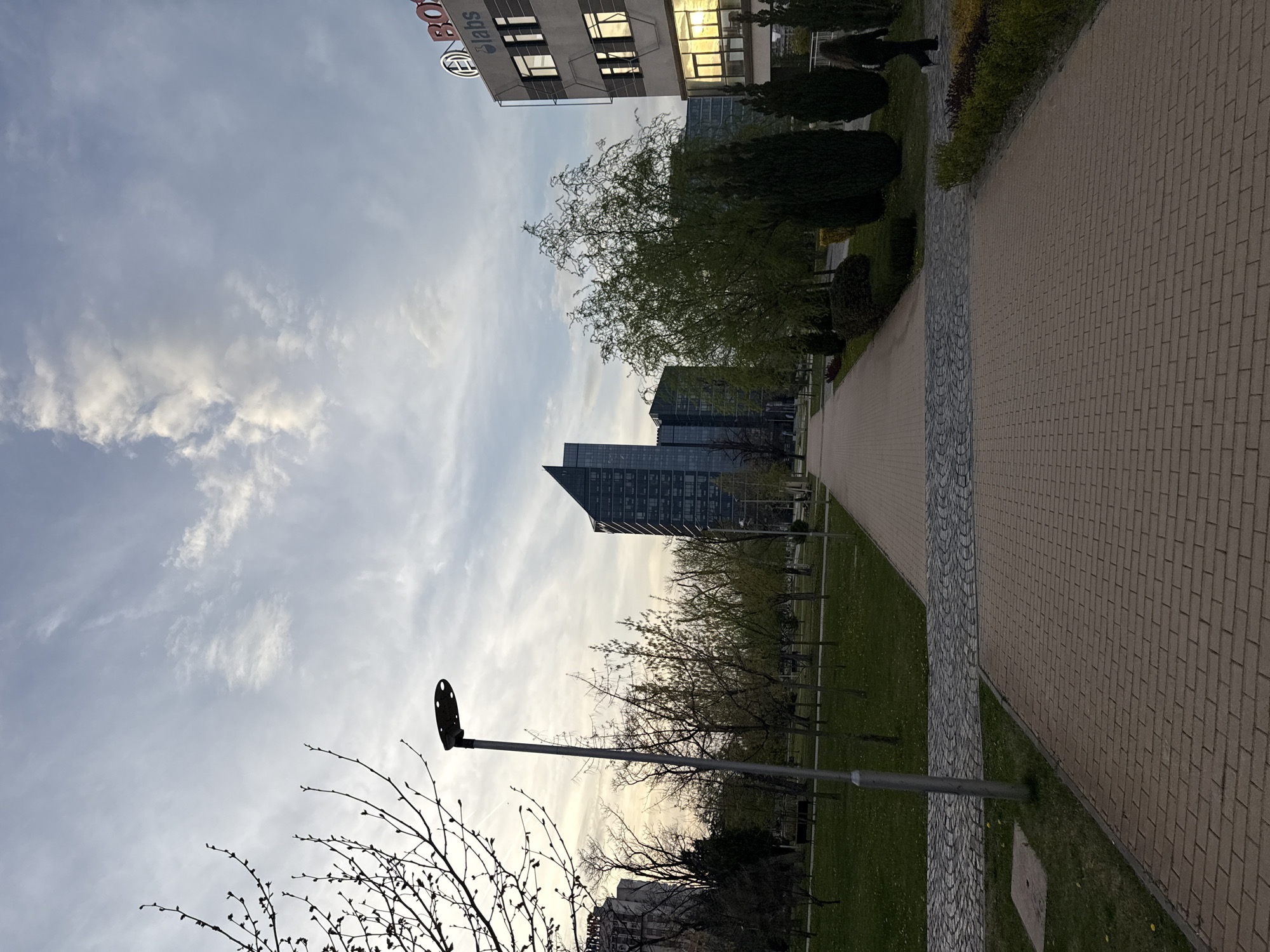 The image captures an urban landscape, likely a business park or campus, depicting modern architecture, green spaces, and a pathway under a partly cloudy sky.
The scene features several modern buildings. Prominently, a light-colored building in the upper right displays the red "BOSCH" logo and the word "labs" in black lettering. Its windows reflect a warm, golden light, suggesting a low sun. Further back, on the left side of the frame, another tall, dark, glass-faced building with a distinctive angular roofline stands out against the sky.
In the foreground and mid-ground, a paved pathway composed of both brick and cobblestone sections extends diagonally from the lower right towards the background. To the left of this path, a grassy area is dotted with trees. Some trees appear bare with budding branches, indicating early spring, while others have already developed lush green foliage. A tall, gray lamppost is visible in the mid-ground on the left, silhouetted against the sky.
The sky is partly cloudy, with patches of bright white clouds and areas of lighter blue, suggesting the transition of day. The warm reflections on the building windows and the soft light imply either late afternoon or early evening. The overall atmosphere appears calm and peaceful within an organized urban environment. No people or explicit activities are visible, presenting a static view of the location. The location context indicates this scene is in Sofia, Bulgaria.