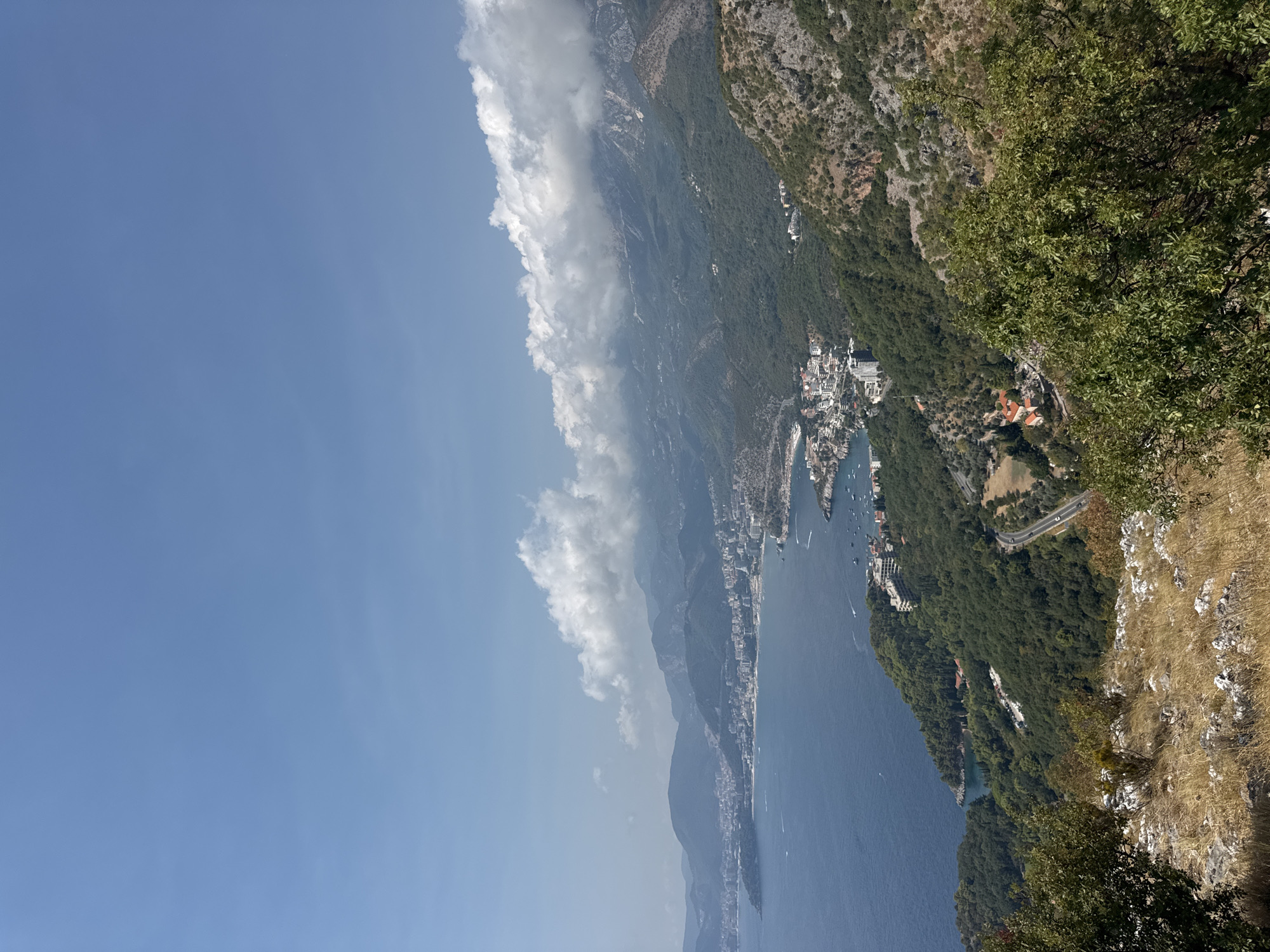 This is a panoramic, high-angle shot of Sveti Stefan, Montenegro, taken on a clear, sunny day with scattered clouds. The image captures a sweeping view of the Adriatic Sea meeting the mountainous coastline. In the foreground, dry, grassy terrain with scattered rocks and bushes suggests a natural, elevated viewpoint.
The middle ground reveals a lush, green, tree-covered hillside that slopes down towards the sea. A winding road is visible, with a few scattered buildings nestled among the trees, including some with red-tiled roofs.
Further out, along the coastline, a developed town or city stretches along the water's edge. Numerous white buildings, many appearing to be hotels or apartment complexes, are clustered together. Boats are scattered in the bay, some closer to shore and others further out. The water is a deep blue, with white wakes indicating the movement of boats.
In the background, a series of imposing mountains rise from the coast. The peaks of these mountains are partially obscured by a dramatic band of white, fluffy clouds that hug the ridgelines. The sky above is a bright, clear blue. The overall impression is one of a beautiful coastal landscape with a significant human presence in the form of urban development and infrastructure.