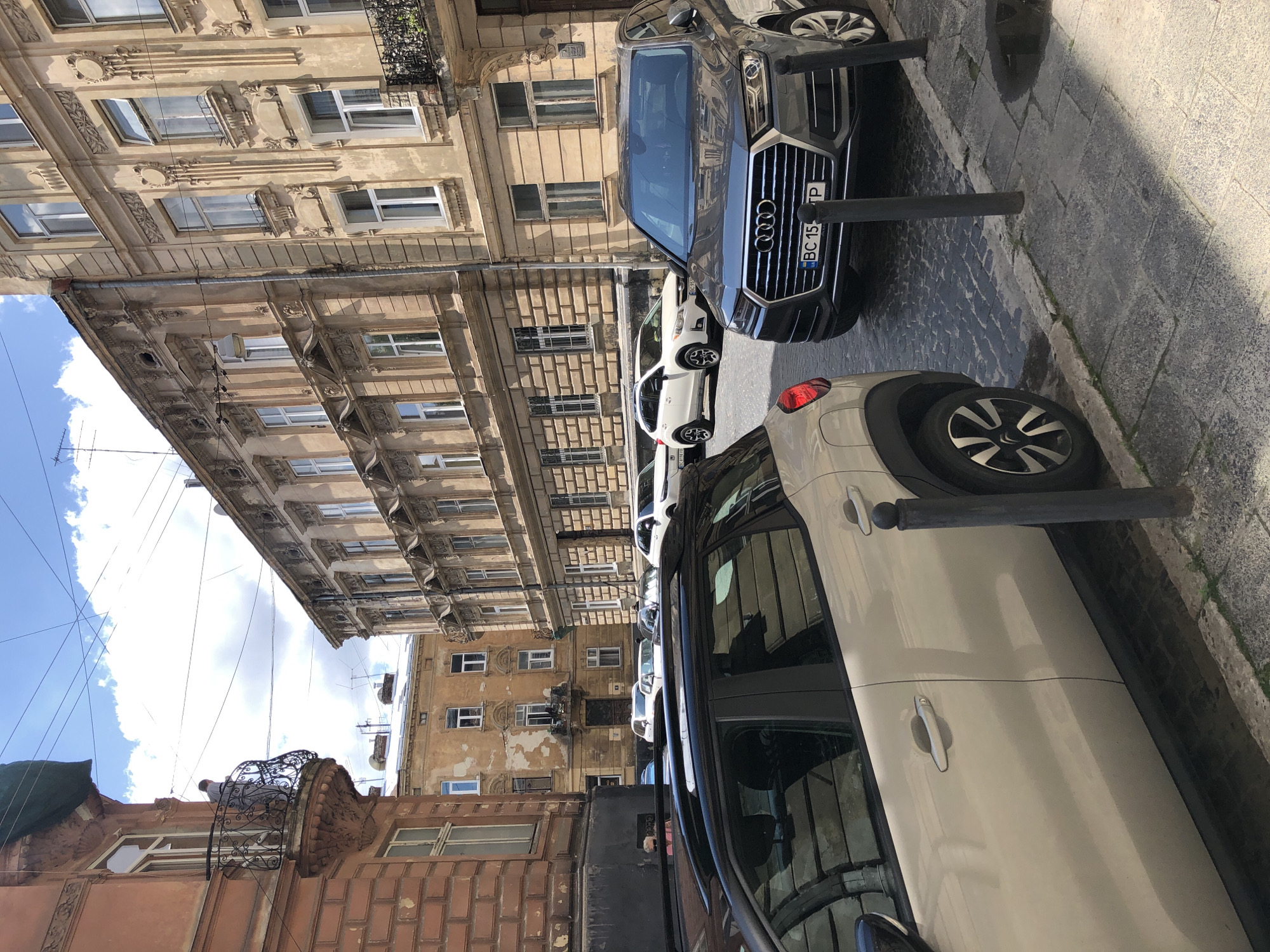 The image shows a street scene in Lviv, Ukraine, on a sunny day with scattered clouds. The foreground is dominated by the side view of a light-colored SUV parked on a cobblestone street. To the right, a dark gray Audi SUV is also parked, displaying a license plate that reads "BC 155 TP".
The background features a row of historic buildings with ornate architecture, characteristic of Lviv's old town. These buildings are several stories high, with numerous windows and decorative detailing on their facades. One building on the left has a reddish-brown brick exterior and a balcony with an iron railing, where a person in a gray shirt is standing. Another building in the middle distance appears weathered and discolored. The street is lined with black bollards. The overall impression is a street in a European city, likely with a historical center.