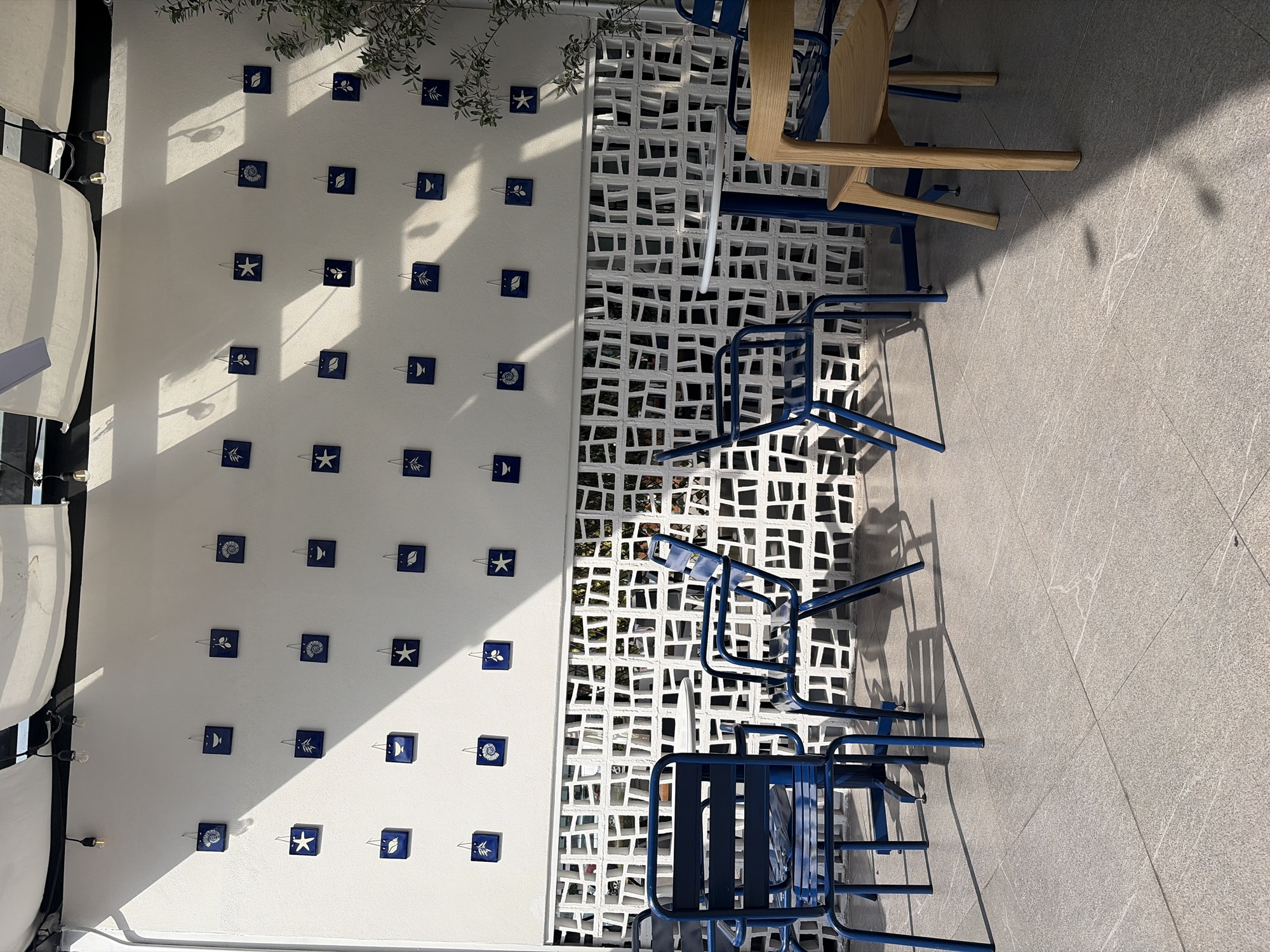 The image depicts an outdoor seating area next to a white wall decorated with small, square, blue tiles featuring marine life designs. A white lattice partition separates the seating area from a garden. Various blue metal chairs and wooden chairs are arranged near the wall and partition. The ground is paved with grey tiles. Shadows from the sun and objects in the image create a contrast on the surfaces. The location is in an unspecified city, Unknown, Unknown.