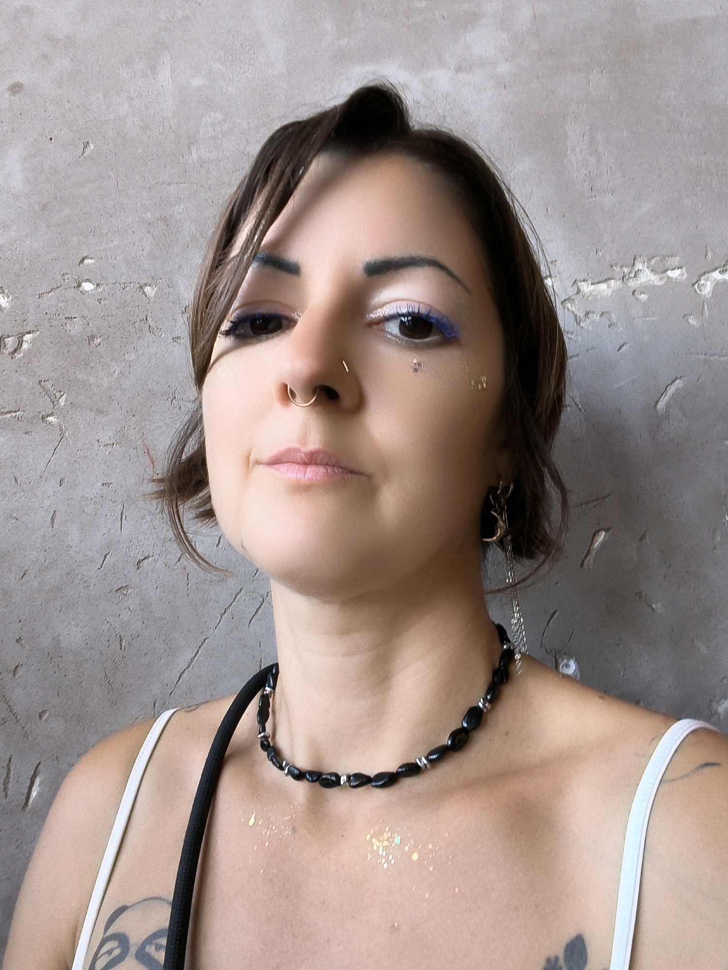 A woman with brown hair, wearing a white tank top and a black necklace, stands against a gray, textured wall. She has a nose ring and is wearing blue eyeliner with glitter accents on her face. Her left shoulder and upper chest are visible, with tattoos on her left arm and chest. The background appears to be a weathered, concrete wall.