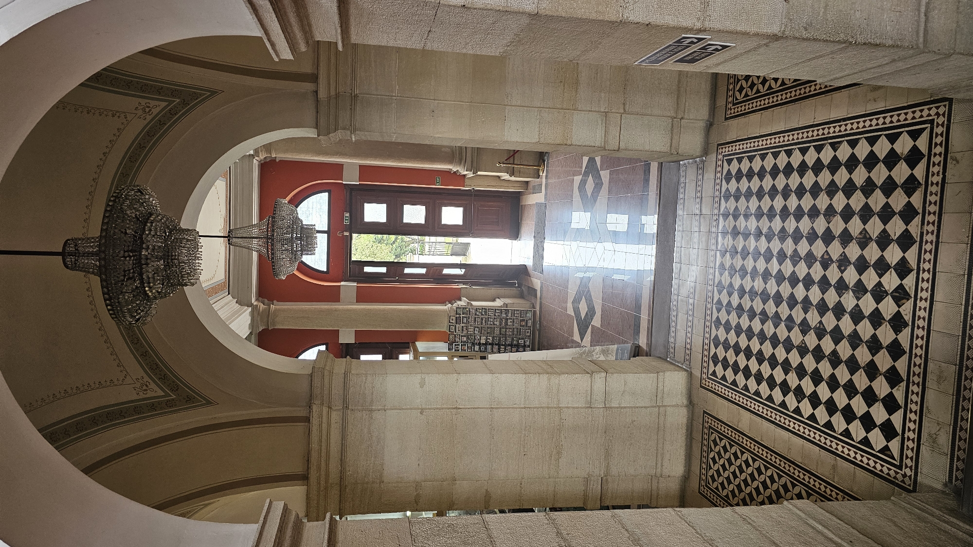 This image depicts the interior of a building, likely a museum or historical site, in Varna, Bulgaria. The architecture features a large arched entryway adorned with a decorative chandelier. A second archway, painted in an orange hue, leads to a wooden door. The floor is tiled with a checkerboard pattern of black and beige tiles. Light enters through the door, creating a reflection on the polished floor. There are no people visible in the scene. A cabinet filled with small items, possibly artifacts or displays, is located beneath the orange archway. The walls and pillars are constructed of stone, showcasing the building's architectural style. The overall impression is one of historical elegance.