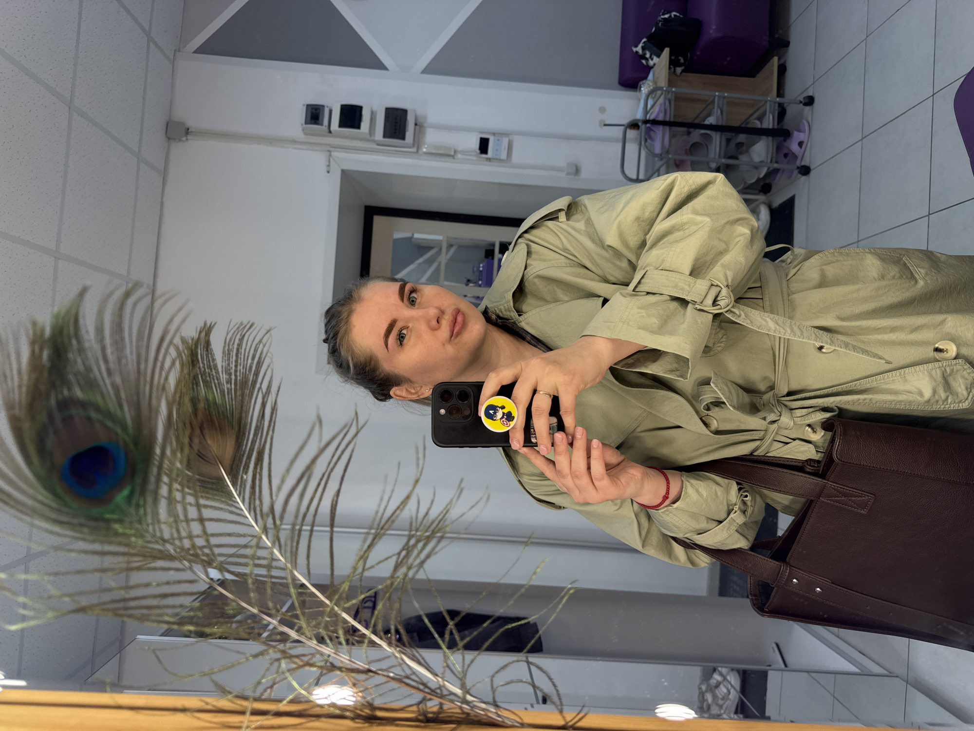A woman is taking a selfie in a mirror. She is wearing a tan trench coat and holding a black smartphone with a yellow cartoon character sticker on the back. She is wearing a red bracelet on her left wrist. A brown leather tote bag is visible. Reflected in the mirror are peacock feathers in a vase. The background shows a white ceiling, a doorway leading to another room, and a purple storage cart with shoes. The photo appears to be taken indoors. The location could be Perm, Russia, although it's hard to confirm this only from the image.