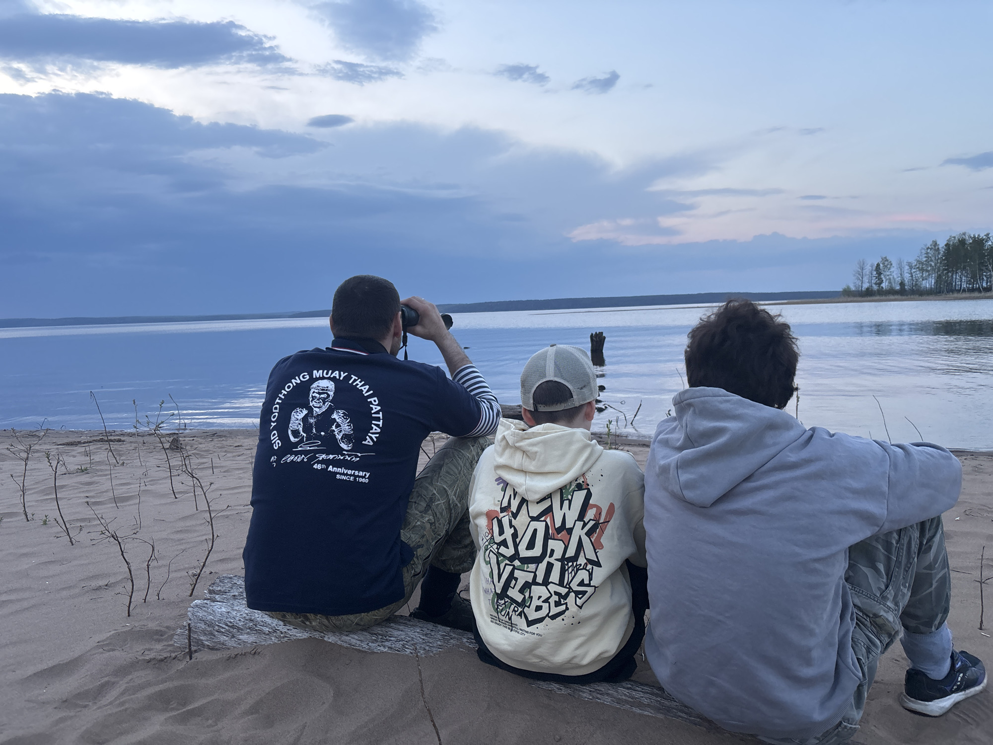 Three people sit on a log at the sandy edge of a body of water, possibly a lake or bay, in Vekoshinka, Russia. The person on the left, seen from the back, is using binoculars and wearing a dark blue t-shirt with a light-colored graphic and camouflage pants. A child in the middle wears a light-colored hoodie with the words "NEW YORK VIBES" on the back and a baseball cap. The person on the right wears a gray hoodie and camouflage pants. The body of water extends to the horizon with some trees visible in the distance. The sky is cloudy with shades of blue and light pink.