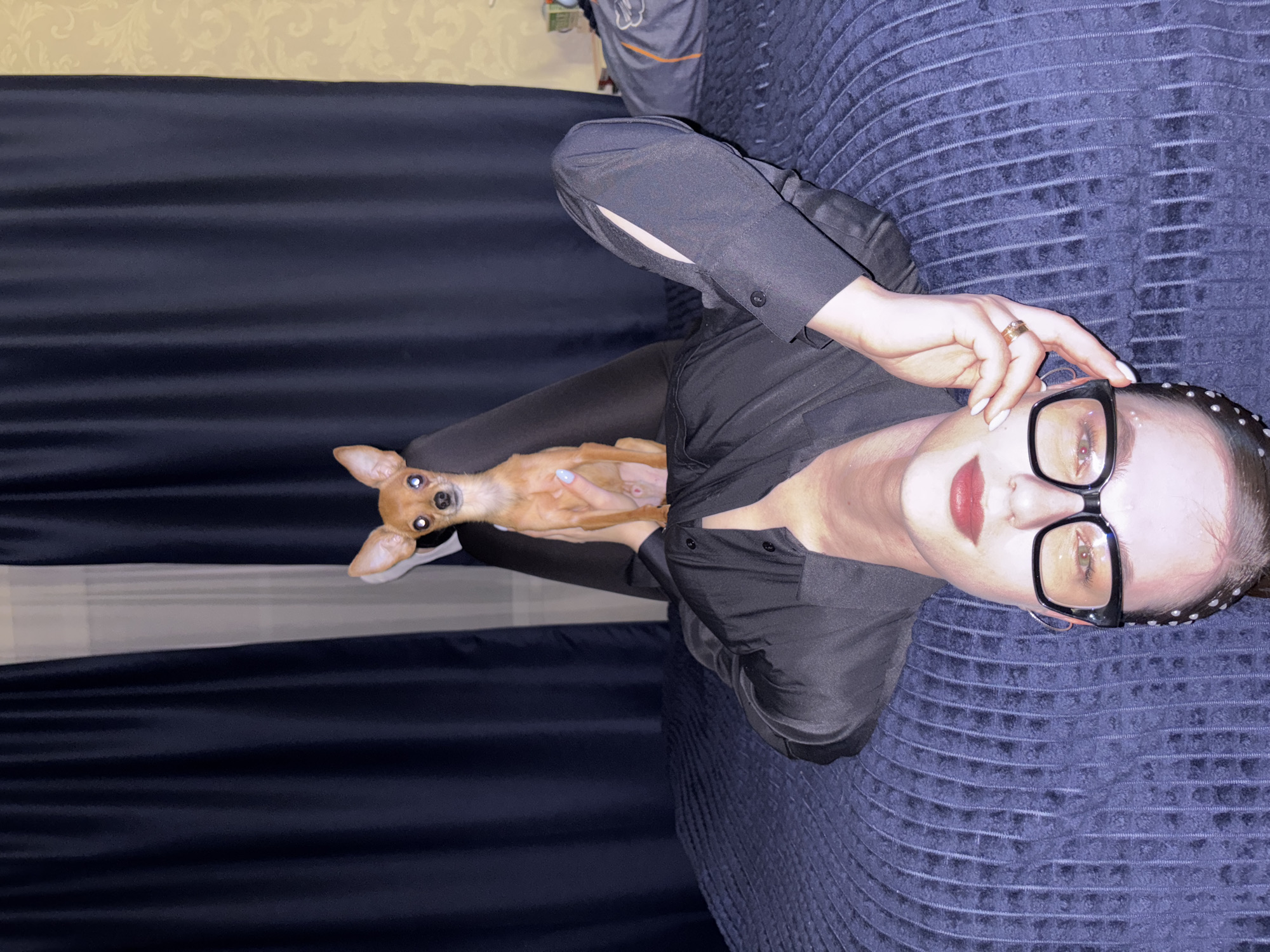 A woman with fair skin, wearing black-framed glasses, a black shirt, and a beaded headband, is holding a small brown Chihuahua dog. The woman is partially reclined on a blue textured surface, possibly a bed or couch. The background includes a dark blue fabric backdrop and a patterned wall. The location is Chaikovsky, Russia.
