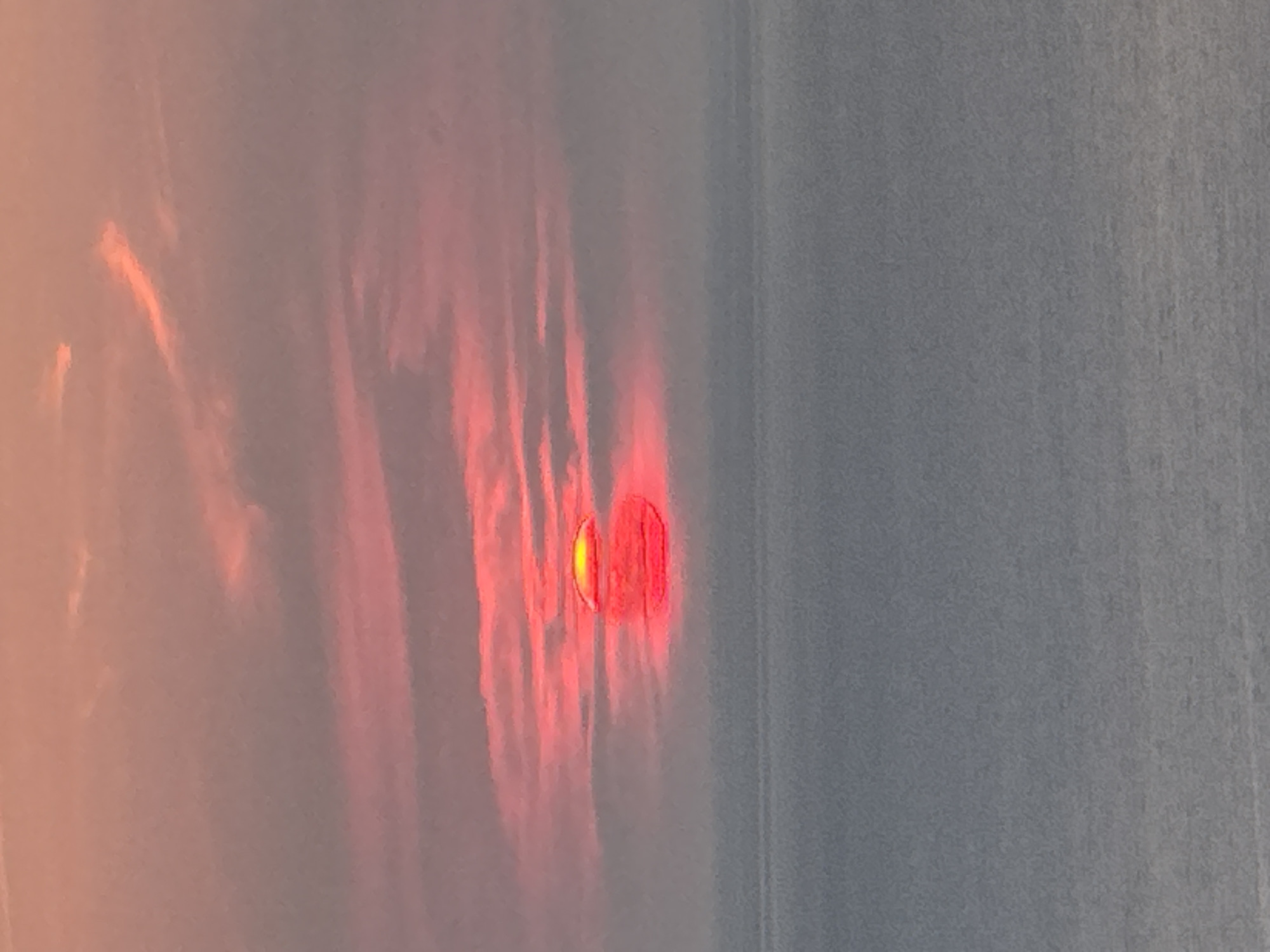A wide shot captures a sunset over the sea, with the sun partially obscured by a hazy horizon. The sky is a gradient of soft oranges and pinks, with streaks of darker clouds suggesting movement. The sun itself is a vibrant red-orange disc, with a brighter yellow glow at its center. Below the horizon, the sea is a muted, grayish-blue, with subtle waves suggesting calm conditions. The scene appears to be a natural landscape, devoid of any man-made structures or human presence, consistent with a coastal location such as Zagora, Montenegro.