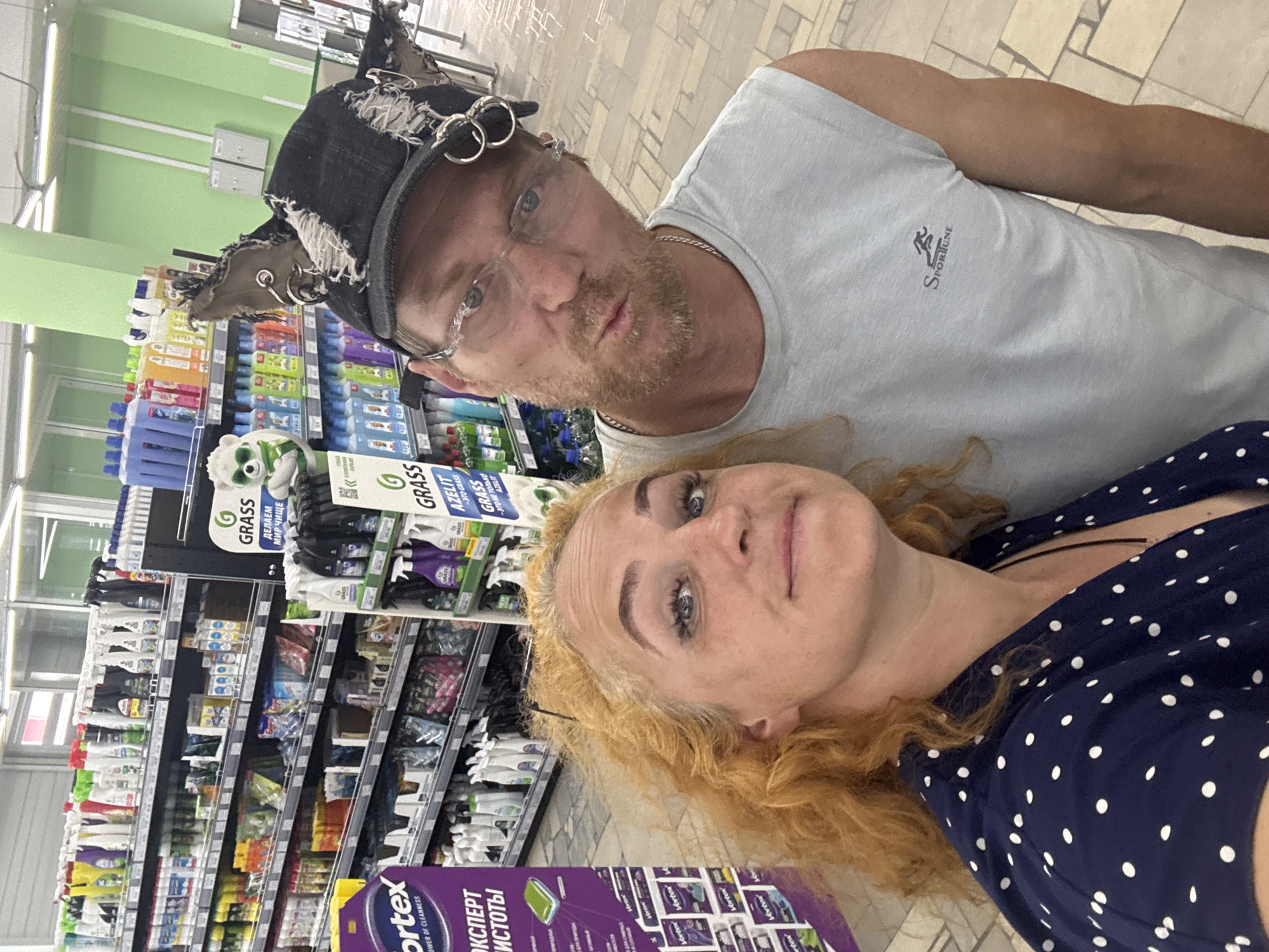 A medium shot from a high angle shows a white man and a white woman inside a retail store. The man is wearing a light blue, sleeveless t-shirt and a black denim cap adorned with piercings and torn fabric. He has a beard and wears clear, rimless glasses. The woman is wearing a dark blue, polka-dot blouse and has blonde, curly hair. She is closer to the camera and her face is partially obscured. Behind them are shelves stocked with cleaning products and other retail goods. The overall lighting suggests an indoor environment. The location is identified as a retail store in Chaikovsky, Russia.