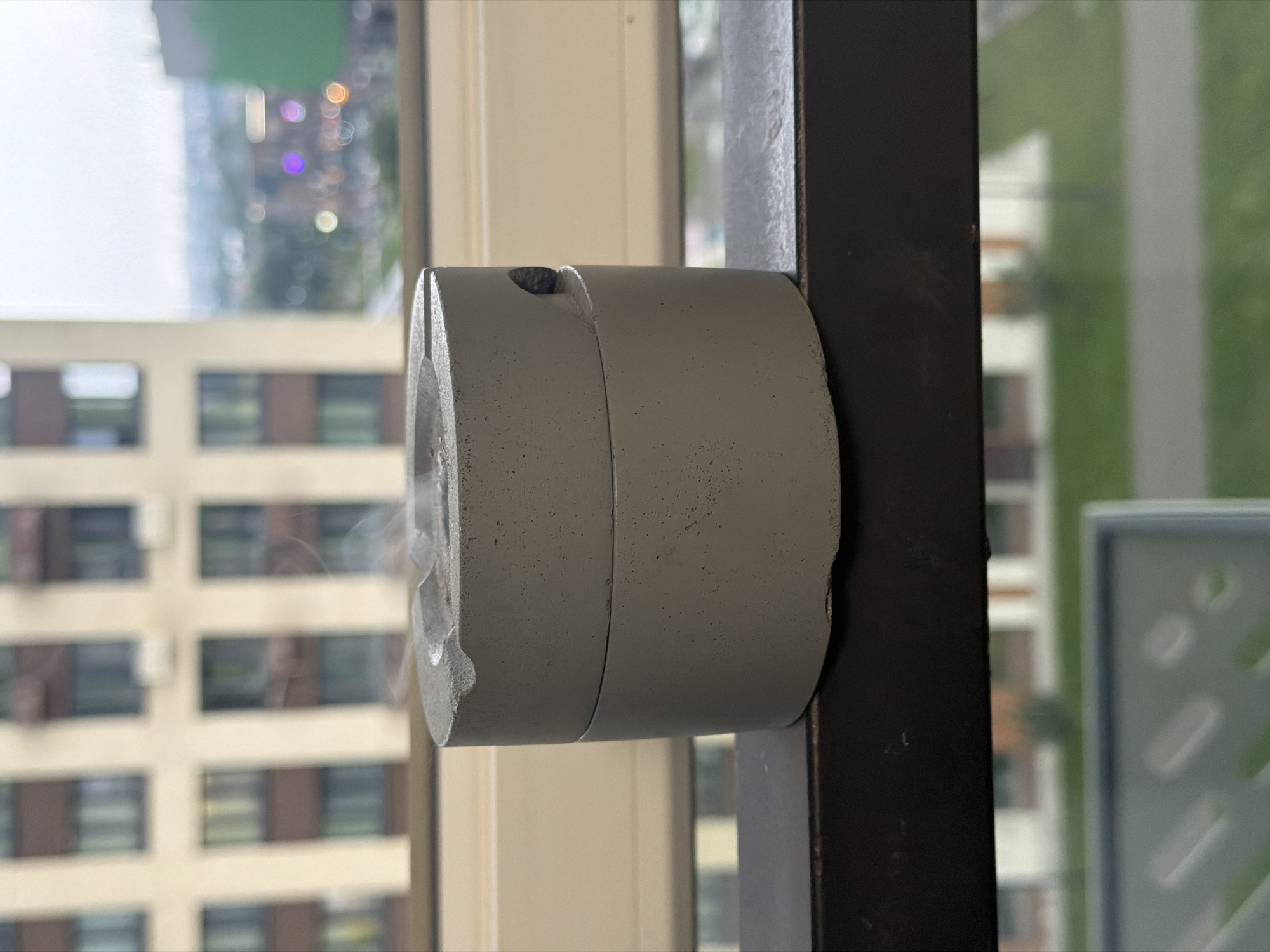 The image shows a concrete ashtray on a balcony railing in front of a window. A building with many windows and balconies is visible in the background through the window, as is a blurred city skyline with some greenery. The ashtray has a small amount of ash in it and a faint wisp of smoke rising from it, suggesting it has recently been used. The lighting suggests it is daytime, possibly overcast. There are no people visible in the image. The location is stated to be in Kazan, Russia.