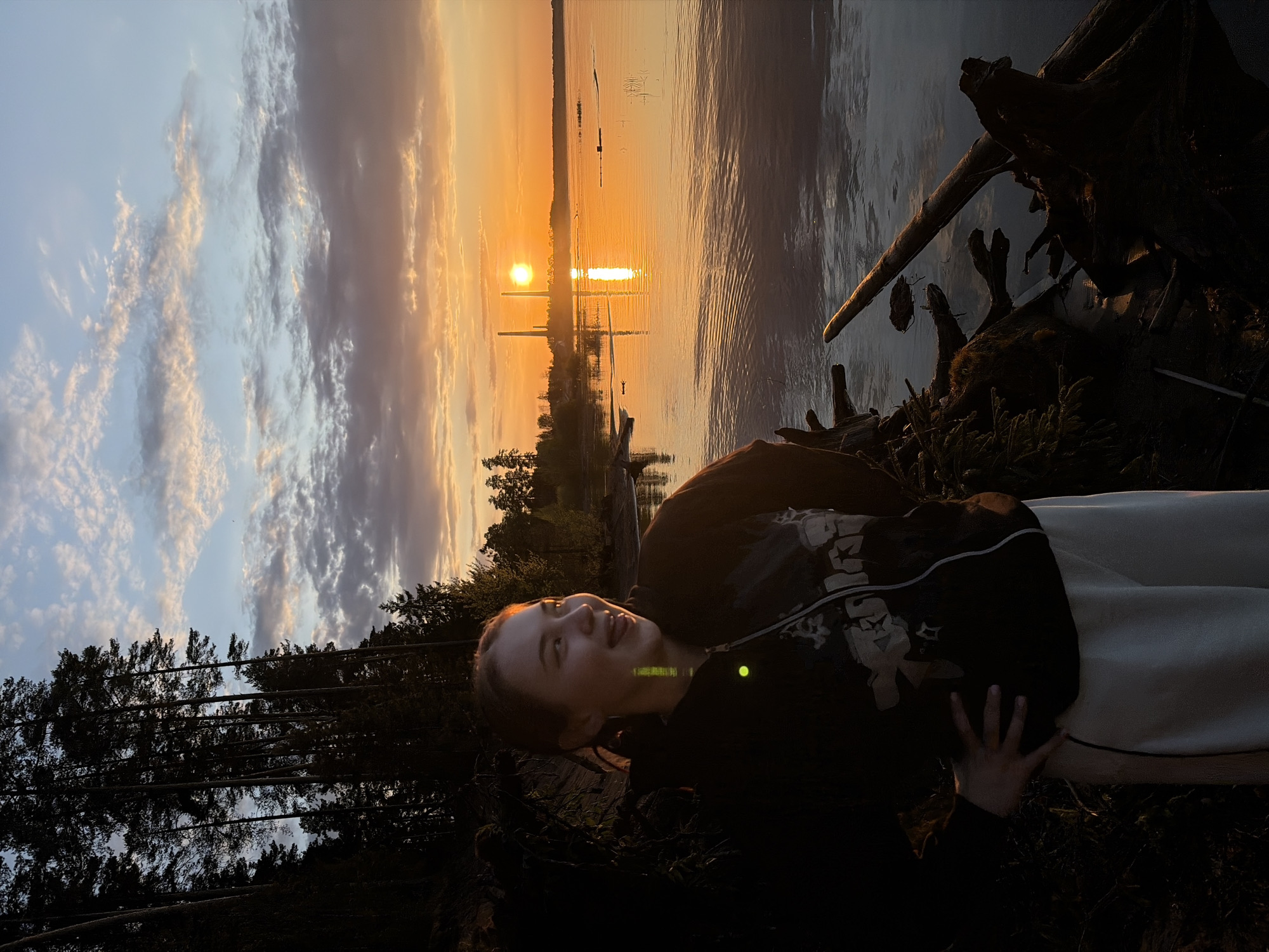 A landscape photograph, rotated 90 degrees counterclockwise, features a young person lying on a grassy area along a shoreline at sunset. The setting sun is directly ahead, casting a bright reflection on the water surface, with tree-covered areas visible along the opposite shore. A dark patch of forest trees borders the lower left of the frame, while driftwood and vegetation occupy the upper right foreground. The young person is dressed in dark clothing with light-colored trousers, turned towards the water with an upward gaze. The city of Chaikovsky, Russia, is not identifiable from this image.