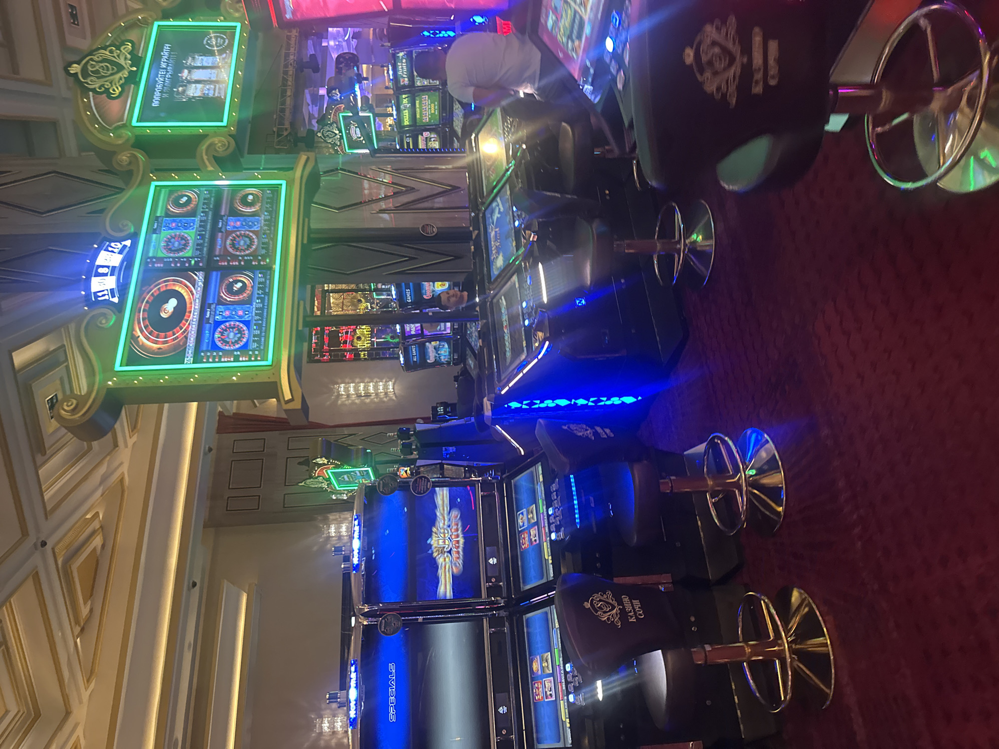 This is a vertical, medium shot of a section of a casino, likely located in Estosadok, Russia, as indicated by the text "КАЗИНО СОЧИ" (Casino Sochi) visible on the chairs. The scene is dominated by rows of brightly lit slot machines and their accompanying bar stools. The slot machines themselves feature multiple screens displaying colorful graphics and game interfaces, with prominent blue and green lighting accents. One of the machines has a roulette wheel simulation.
There are at least two people visible, though not clearly depicted. One appears to be a man in a white t-shirt standing behind some machines, and another man is partially visible in the lower left section of the image, looking towards the machines. The overall atmosphere is one of vibrant entertainment and gaming. The red carpet flooring is visible, adding to the luxurious feel of the casino. The lighting suggests it is nighttime or an indoor environment with artificial illumination. Some machines display text in Russian, such as "ПОПРОБУЙТЕ! ИГРАЙТЕ И ЭМИГРЫВАЙТЕ" which translates to "Try! Play and Win." and "ALL GAMES". The decorative elements around the machines, with gold accents and ornate designs, suggest a high-end establishment.