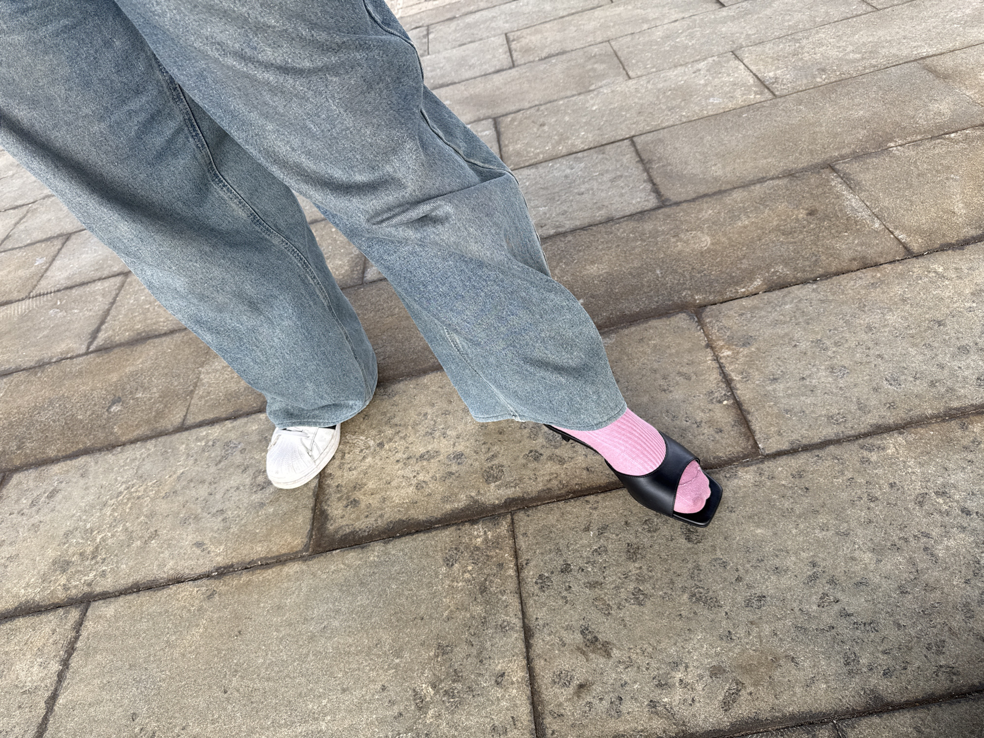 The image shows a person's legs and feet walking on a paved outdoor surface. The person is wearing loose-fitting light blue jeans. On the left foot, a white sneaker is visible. On the right foot, a black slide sandal is worn with bright pink socks. The ground is made of large, rectangular stone slabs. The lighting suggests it is daytime, possibly outdoors in a city. There is no visible indication of the specific location within Vnukovo, Russia.