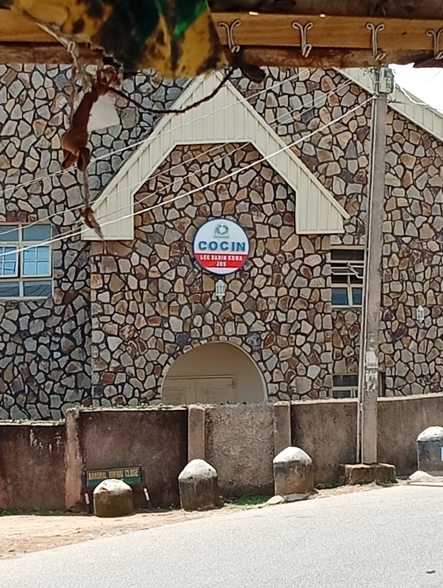 The image shows the exterior of a building with a stone facade in Jos, Nigeria. A circular sign with the text "COCIN LCC BADIRI KOWA JOS" is prominently displayed on the building. Below the building, a low concrete wall with a sign reading "BANSAL BINDA CLOSE" is visible. Two cylindrical stone bollards are positioned in front of the wall. The environment appears to be outdoors, with a paved road in the foreground and what seems to be a street or driveway. The building has multiple windows, and some overhead elements suggest a covered entrance or overhang. The overall impression is of a public or religious institution.