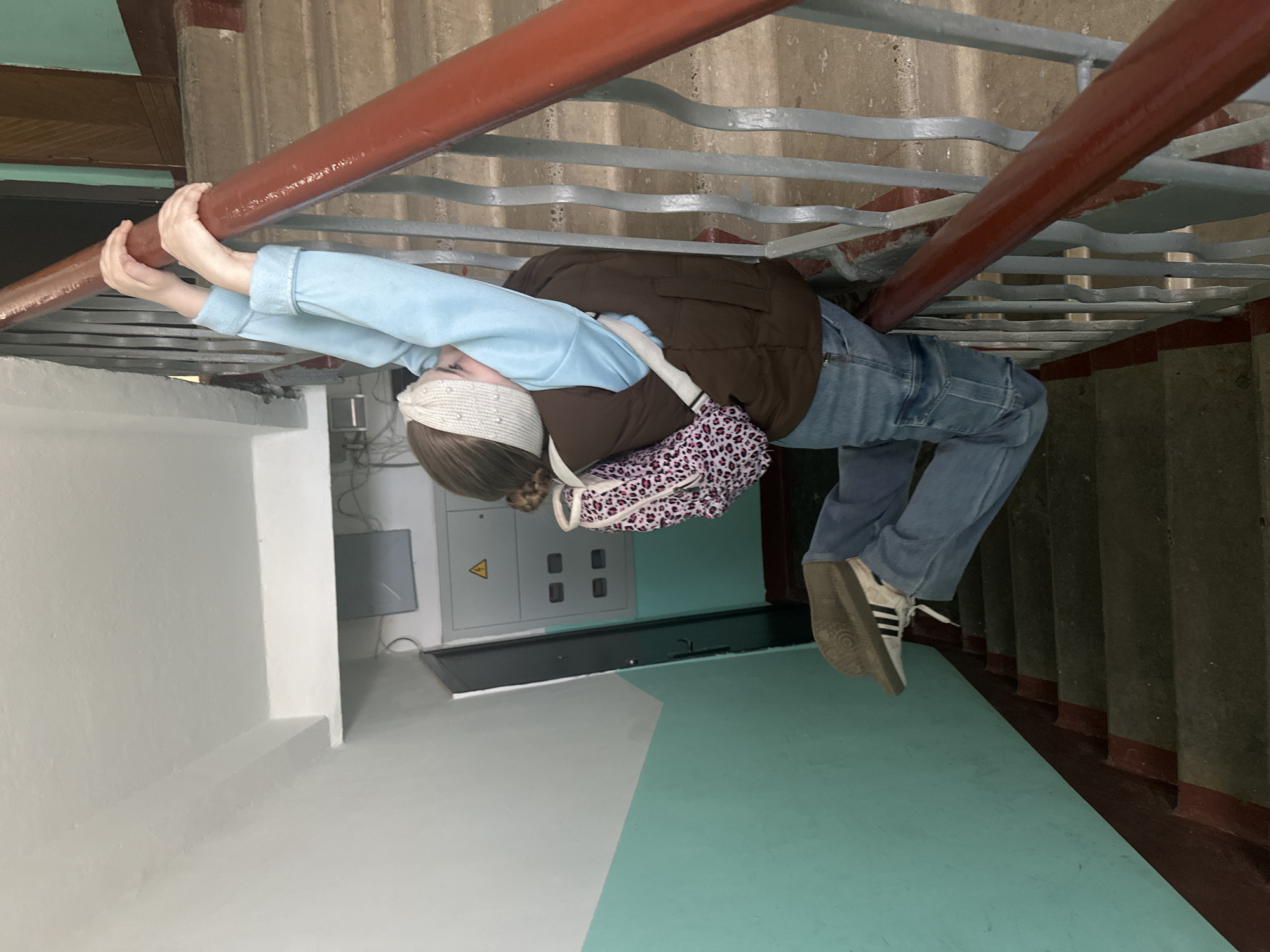 A person is hanging onto a red railing in what appears to be a stairwell. The person wears a light blue shirt, jeans, a brown vest, and Adidas sneakers. A leopard print purse hangs from the person's shoulder. The person's eyes are covered by a beaded headband. The location may be Chaikovsky, Russia. The stairwell has grey metal railings with a red handrail and painted walls. A utility box is visible on the wall.