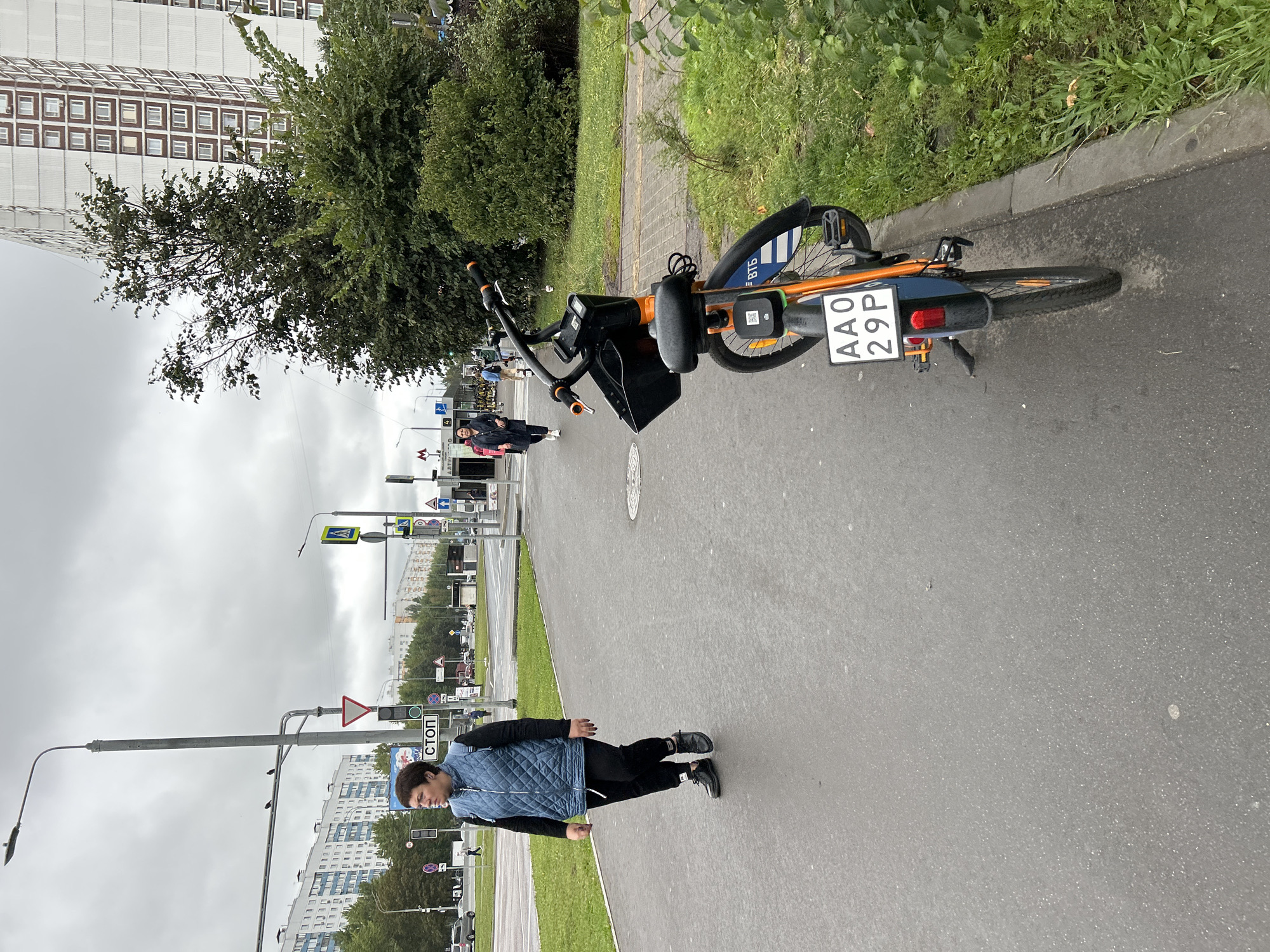 A woman walks along a wet sidewalk in Moscow, Russia. She is wearing a blue quilted vest over a black long-sleeved shirt and black pants, and black shoes. She is looking to her left. In the foreground, an orange and blue rental bicycle is parked. The bicycle has a black basket and a license plate with the characters "AA0 29P". Behind the woman, there are traffic lights, street signs, and a building with many windows. The sky is overcast and the ground is wet, suggesting it has recently rained or is drizzling. The scene appears to be a street in a residential or commercial area of the city.