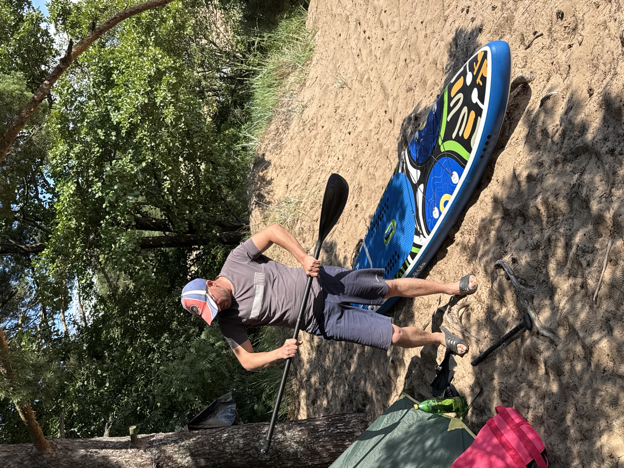 A man wearing a grey t-shirt, blue shorts, a red and blue baseball cap, and grey sandals stands on a sandy embankment. He is holding a black paddle horizontally with both hands, preparing to engage with a blue and white paddleboard that lies on the sand in front of him. To the left, a green tent is pitched, and a pink backpack is placed on the ground. A green bottle rests beside the tent. The scene is set in a wooded area with tall trees and dense green foliage in the background. The sandy ground is littered with pine needles and small twigs. Sunlight filters through the trees, casting dappled shadows on the sand. The location is Zelenaya Roshcha, Russia.