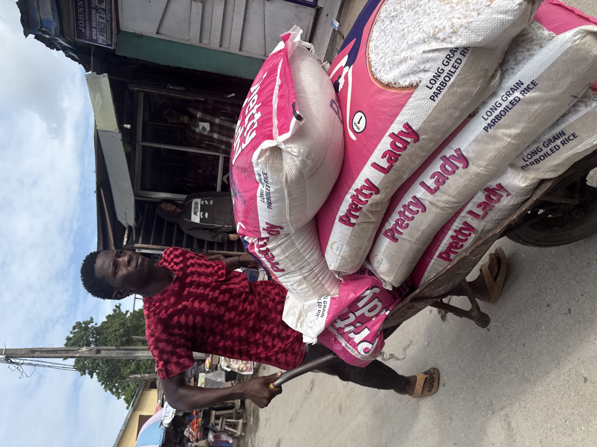A man is depicted pushing a heavily loaded handcart along an outdoor street in Lagos, Nigeria, during what appears to be daytime under an overcast sky. He is wearing a red patterned t-shirt, dark trousers, and sandals, holding the cart's handle with a focused expression, indicating the physical effort involved in transporting the goods.
The handcart is piled high with several large sacks of rice. The sacks are primarily pink and white, prominently displaying the brand "Pretty Lady LONG GRAIN PARBOILED RICE," with some also showing "GRADE 1." One of the pink sacks has an opening or tear at the top, revealing the white rice inside.
In the background, typical urban architecture is visible, including buildings with corrugated metal elements. A sign for "JOHNNY COMMUNICATIONS" is partially visible on one building, advertising "REPAIR UNLOCKING FLASHING" services, along with contact numbers: "08130583103" and "0907978594." A utility pole and some foliage from a tree are also present in the distance. The scene captures a moment of daily commerce and manual labor within a busy street environment, characteristic of a market or commercial area in Lagos.