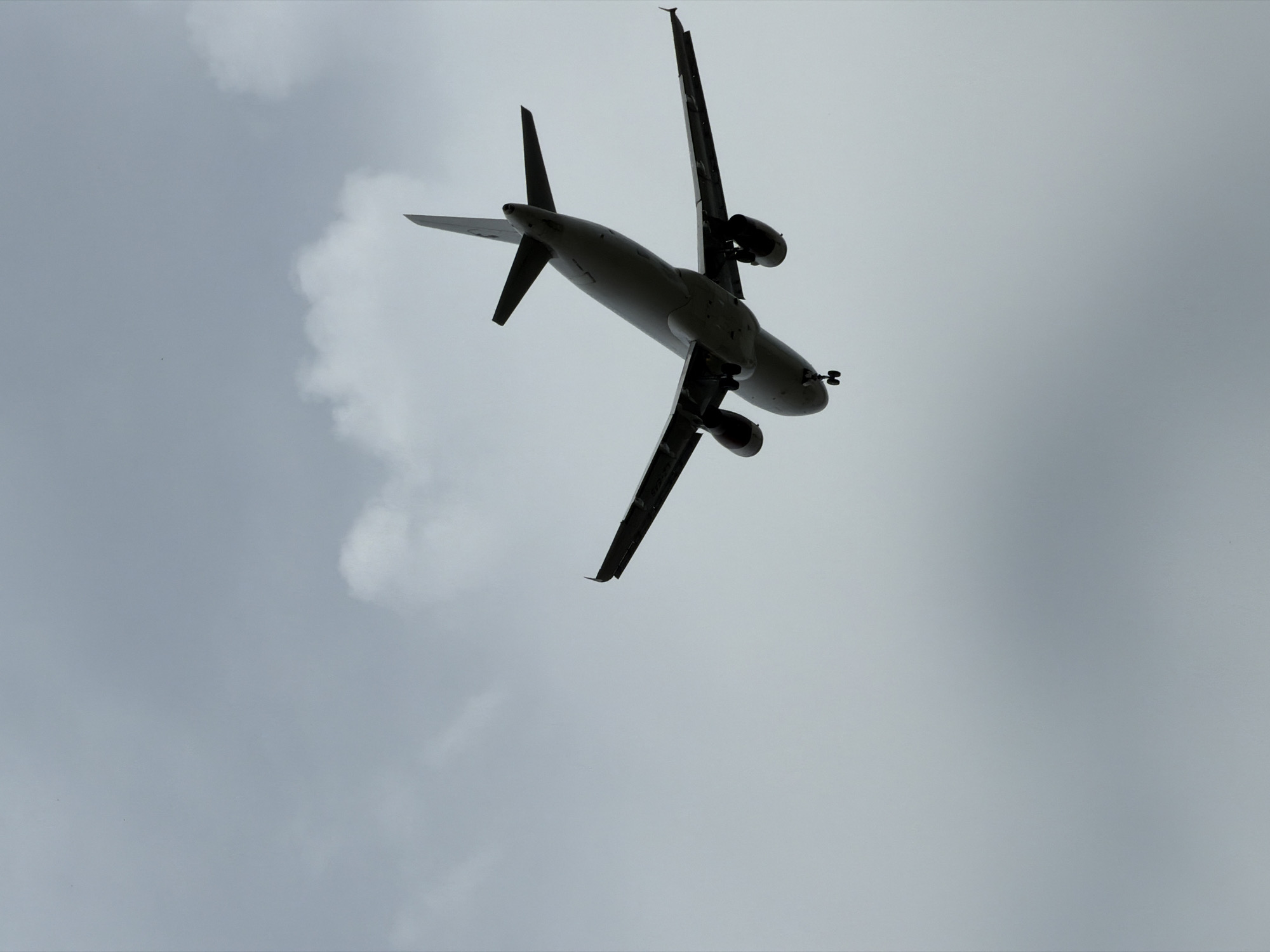 An airplane is captured in silhouette against a cloudy sky, appearing to be in the process of landing or taking off. The underside of the aircraft is visible, revealing its landing gear extended. The scene is underexposed, with the plane rendered as a dark shape against the lighter, overcast sky. There are no discernible landmarks or environmental cues in the image to suggest a specific location within Burgas, Bulgaria. The focus is solely on the airborne aircraft.
