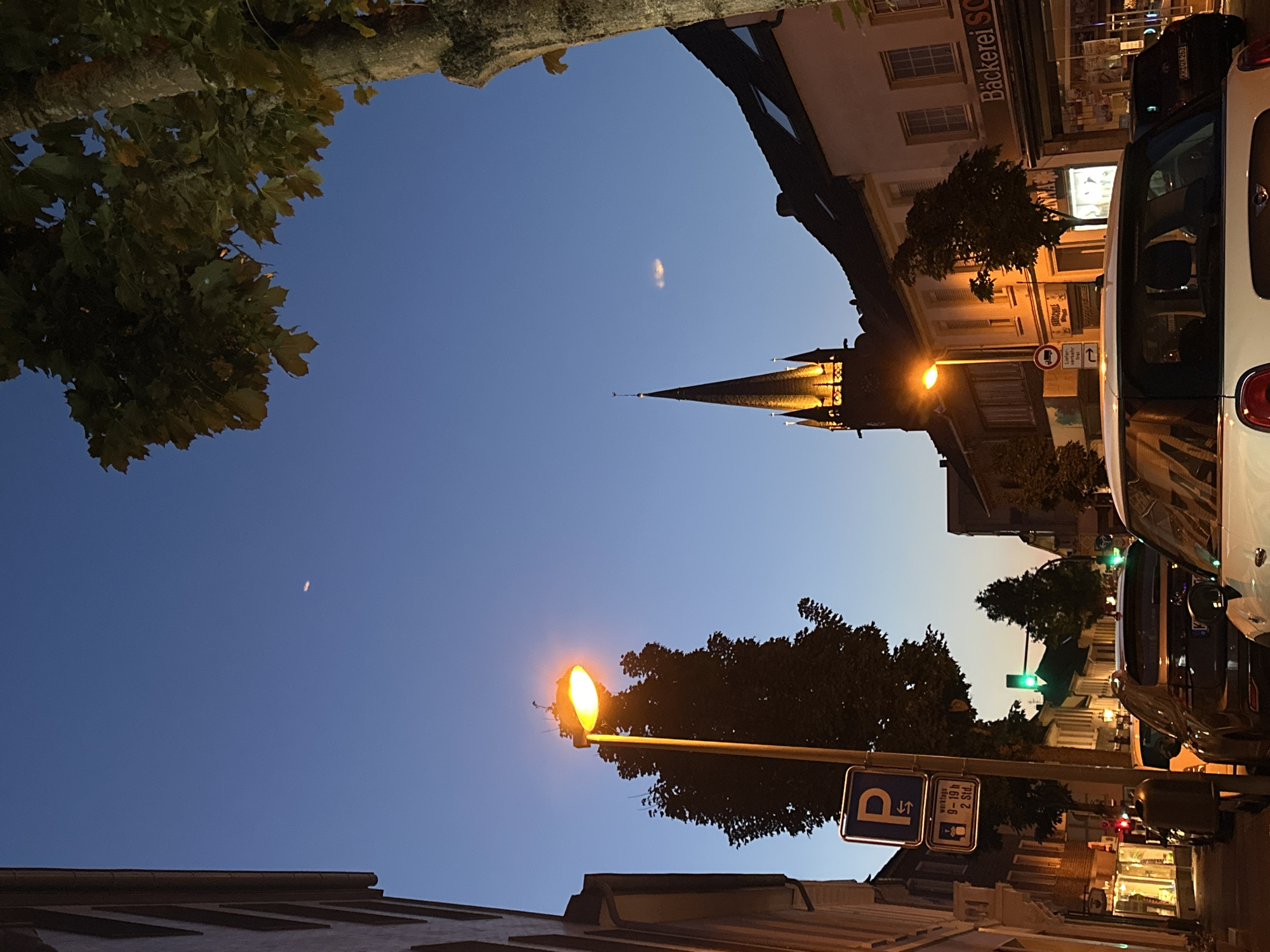The image depicts a street scene in Krefeld, Germany, at dusk. A street lamp illuminates the foreground with its warm yellow light, casting a glow on the surrounding trees and buildings. The sky is a deep blue, with a faint crescent moon visible near the top.
In the background, the spire of a church rises majestically, its architectural details accentuated by the artificial lighting. The street is lined with buildings, some appearing to be shops, with windows displaying goods or illuminated interiors. Cars are parked along the street, indicating a typical urban setting. A traffic light shows a green signal.
The overall impression is one of tranquility and the transition from day to night in a European city. The scene is devoid of people, focusing instead on the architectural elements and the ambient lighting.