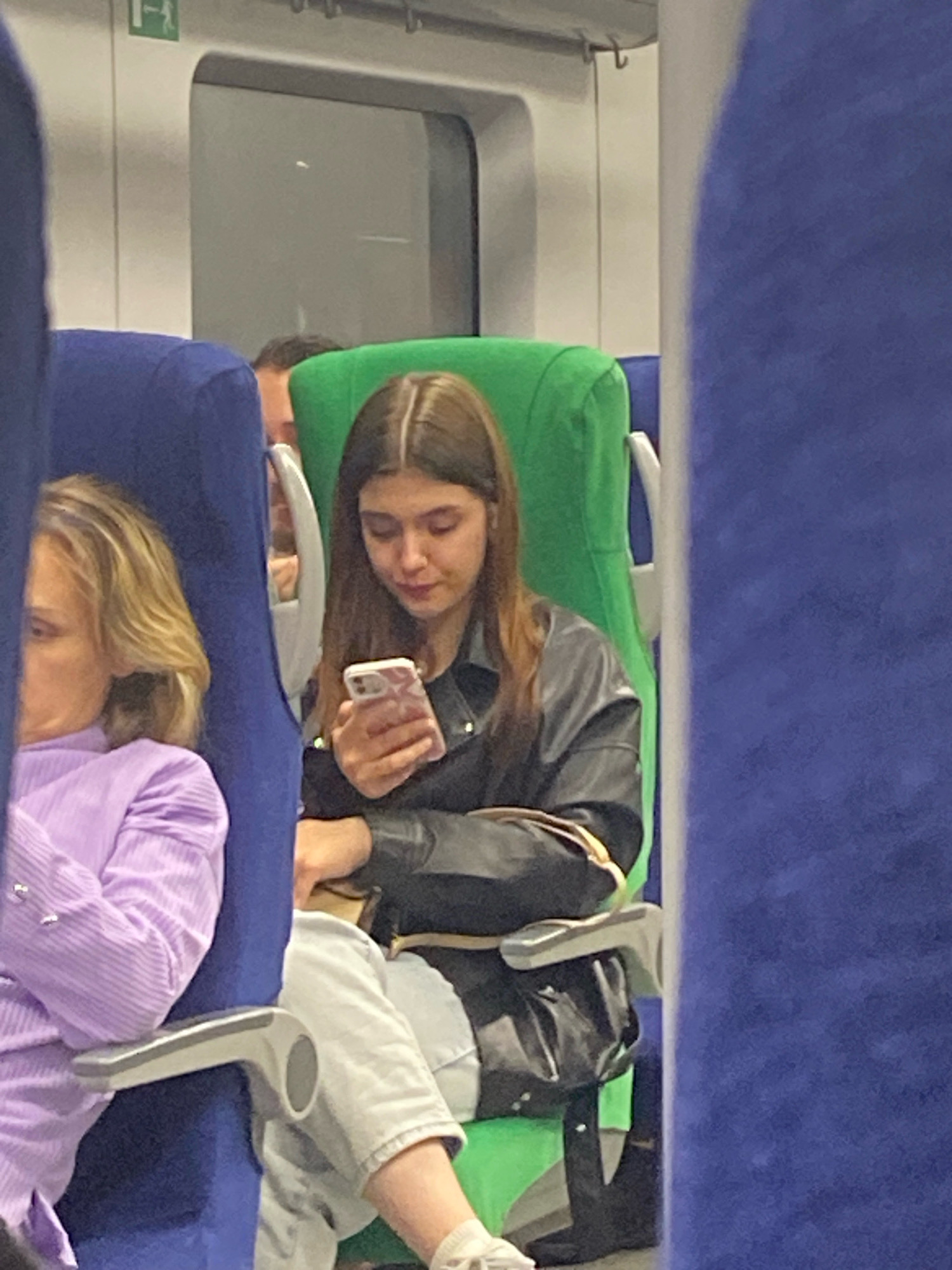 The image shows a young woman with long brown hair looking down at her smartphone. She is seated on a train, indicated by the blue and green seats, and the window with a visible evacuation sign. She is wearing a black leather jacket over a black top. Next to her, a woman with blonde hair is partially visible, wearing a lavender-colored shirt. The woman on the phone is holding the phone in her right hand and appears to be scrolling or typing. A black bag is visible on the seat beside her. The overall environment suggests a public transportation setting, likely a train. The presence of the evacuation sign with Russian text suggests the location is within Russia, possibly Moscow.