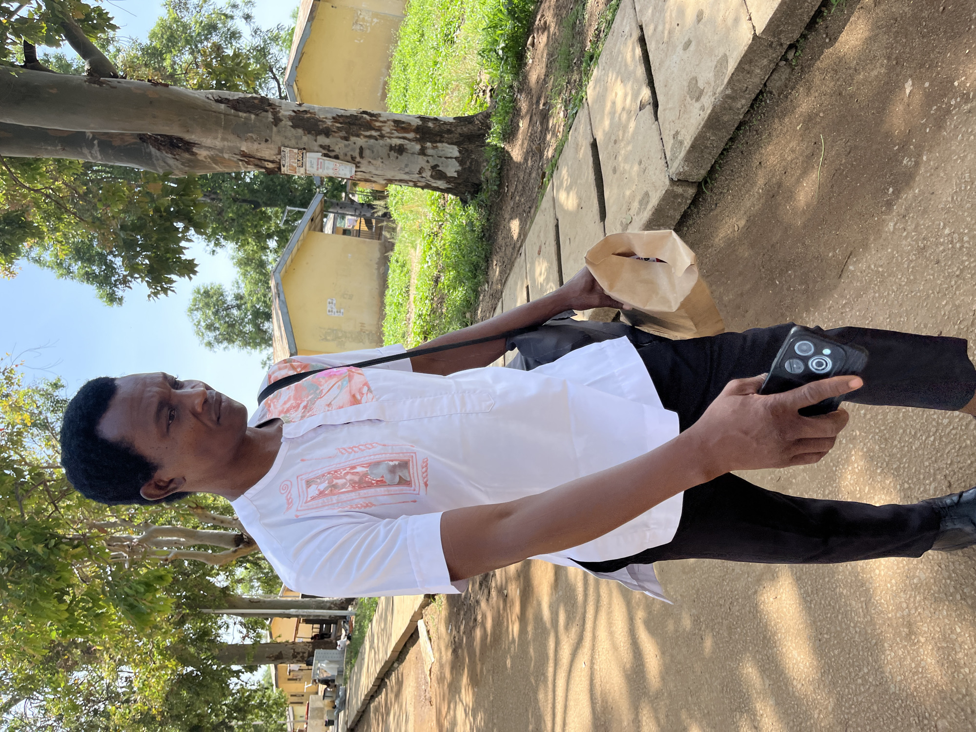 The image shows a man standing outdoors on a sunny day in what appears to be a community or campus setting in Rafin Par, Nigeria. The man, with dark hair and skin, is wearing a white long-sleeved shirt with subtle light orange/pink patterns, black trousers, and dark shoes. He holds a brown paper bag in his left hand and a black smartphone with multiple cameras in his right hand. A black strap or bag is also visible around his shoulder.
He stands on a dirt path bordered by concrete slabs, looking slightly upwards and to his left with a neutral, observant expression. The background features a prominent large tree with light-colored, somewhat peeling bark and green foliage, casting some shadows. Behind this tree and further to the right, several yellowish buildings are visible, interspersed with other trees and green areas. Small, rectangular white posters or signs are affixed to the trunk of the large tree, but their text is illegible. The bright blue sky and the quality of light suggest it is either morning or late afternoon.