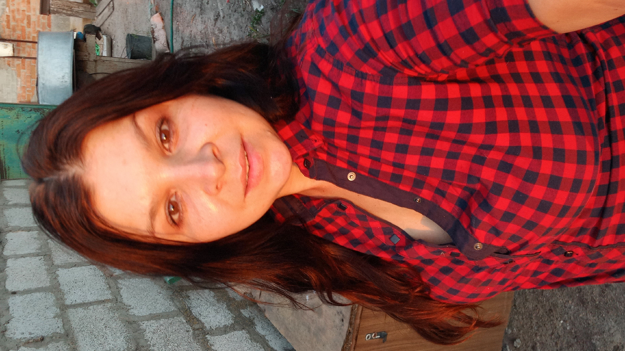 A medium shot photo shows a woman with long, dark hair standing outdoors. She is wearing a red and navy blue plaid button-up shirt. The background consists of a cinder block wall on the left and a rustic outdoor setting with some structures and debris in the background. The lighting suggests it may be sunset or sunrise, casting a warm glow on her face. The location is identified as Klyntsi, Ukraine.