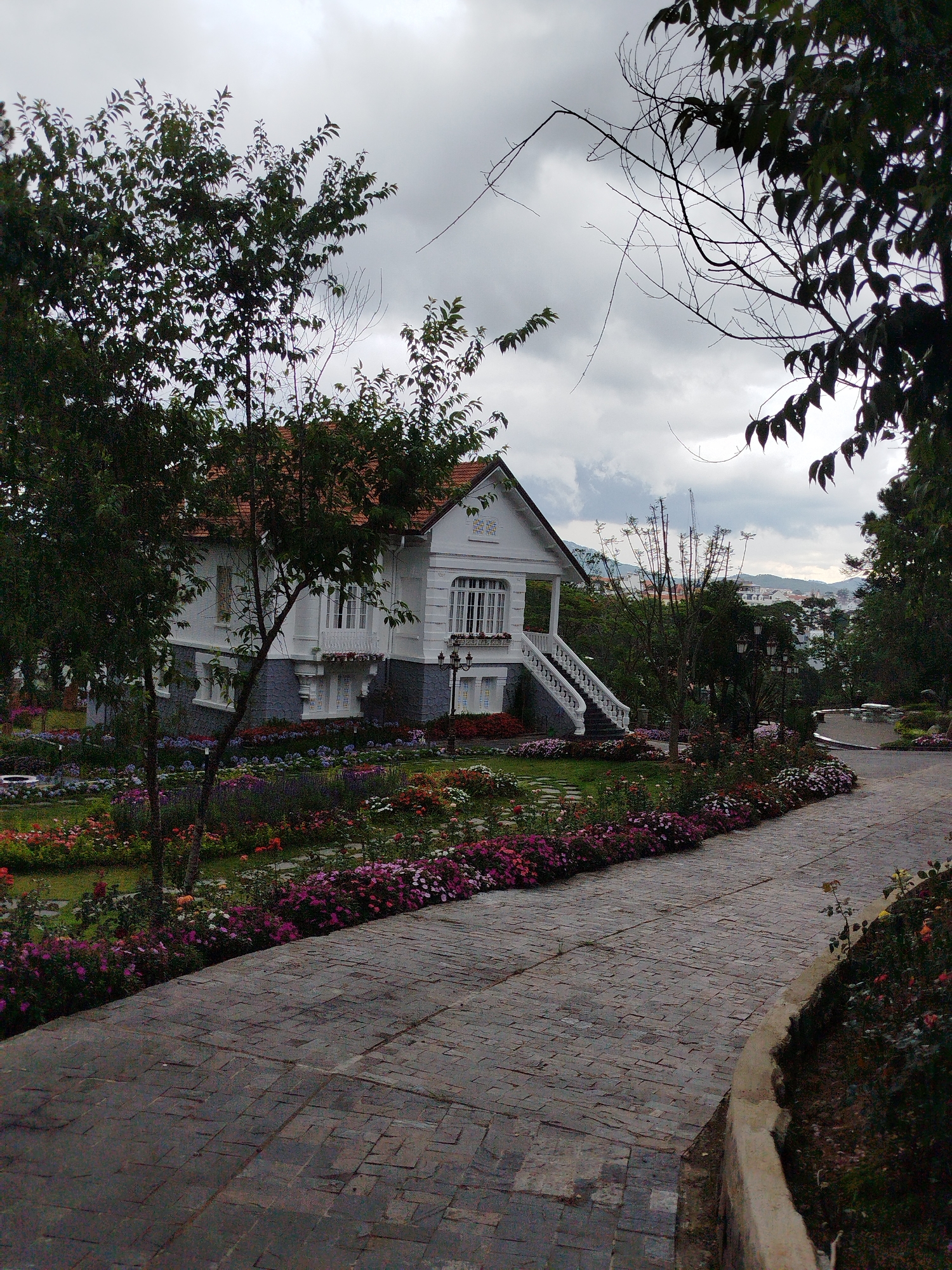 A white house with a red tile roof stands amidst a vibrant flower garden, positioned at the edge of a cobblestone path. The house has two stories. Trees partially obscure the house. A stairway with a white railing leads to the entrance. The cobblestone path is lined with flowerbeds of pink and purple flowers. The sky is overcast. In the background, a city is visible atop hills, indicating the location is in a mountainous region. This scene appears to be within the city of Da Lat, Vietnam, due to the architectural style and the landscape. No people are visible in the image.
