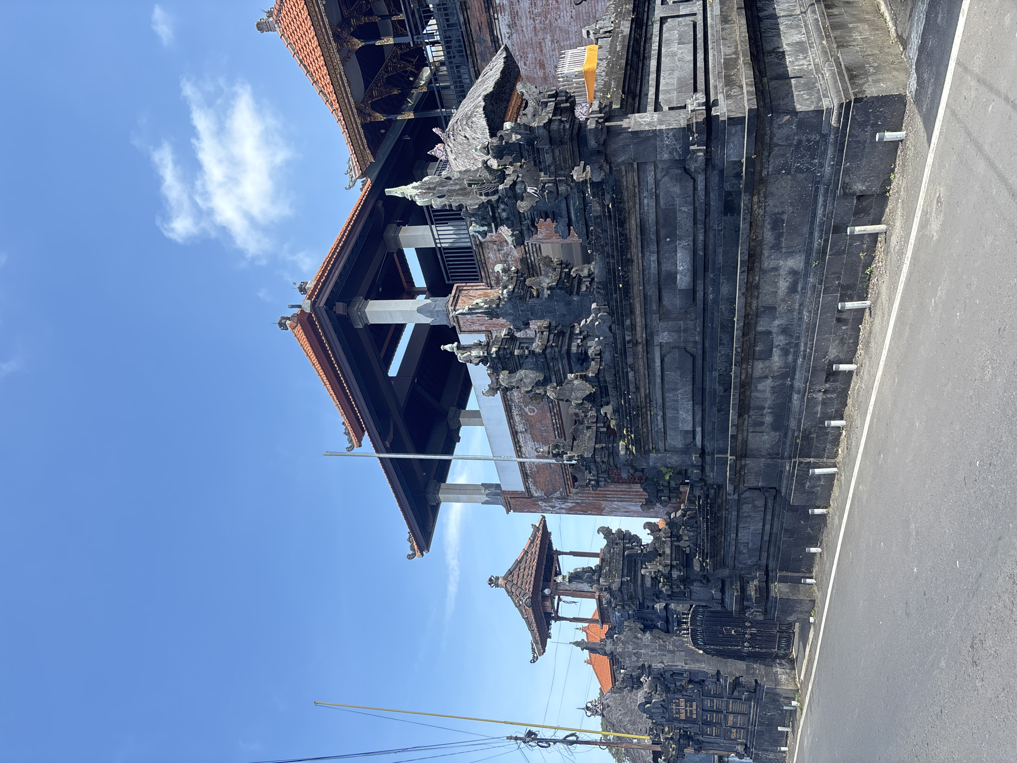 The image shows a traditional Balinese gate located in Kuta Selatan, Indonesia. The gate is constructed of dark grey stone and features intricate carvings and multiple tiers of roofing. The architecture has a prominent layered effect with many ornate, statues, and decorative elements. The gate is positioned on the side of an asphalt road with a white line indicating the edge of the lane. Above, a clear blue sky with scattered white clouds is visible. Power lines run along the left side of the frame.