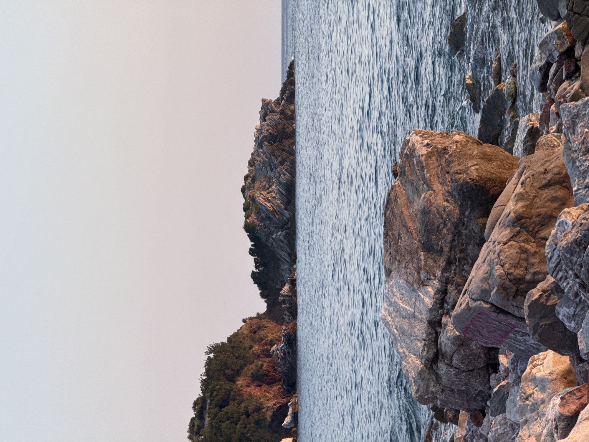 The image depicts a rocky shoreline with the sea on the right side and a cliff with vegetation on the left. The composition is vertical, with the sea and rocks occupying the right half and the cliff and sky the left. The sea has a rippling surface, suggesting a gentle breeze. The rocks are varied in size and texture, with some appearing weathered and others more jagged. The cliff is covered in dense green vegetation, with some brown earth visible. The sky is a pale, hazy grey, indicating either overcast weather or early morning/late evening light. The overall impression is one of a natural, rugged coastal landscape. There is no discernible activity or event taking place. A notable detail is some faint pink graffiti on one of the larger rocks. No people or distinct objects are visible beyond the natural elements of the scene.