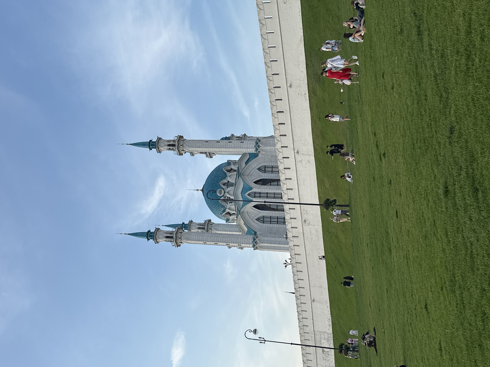 The image captures a view of the Kul Sharif Mosque in Kazan, Russia. The mosque, with its distinctive blue dome and tall white minarets topped with turquoise spires, is situated behind a high, crenelated white wall. The wall runs horizontally across the mid-section of the image, with a steep, grassy incline leading up to it from the foreground. The sky is a clear, light blue with wispy white clouds.
In the foreground and on the grassy slope, various individuals are present. Some are walking, while others are sitting or lounging on the grass. To the left, a lamppost with an ornate lamp stands on the edge of the grassy area. Several people are scattered across the lawn, some in small groups, others individually. A group of people are walking together on the slope, and another group is gathered further up near the wall. A few individuals are seated on the grass in the foreground. The overall scene suggests a pleasant day with people enjoying the public space in front of the iconic mosque.