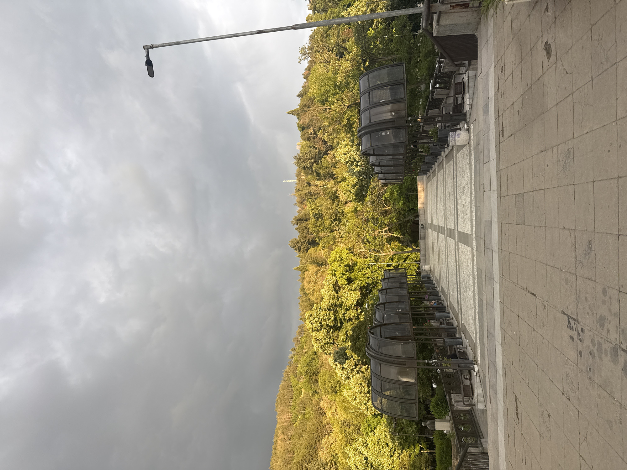 The image shows a paved pedestrian walkway leading away from the viewer, flanked by metal structures resembling covered bus stops. A steep, densely forested hillside rises on the left side of the walkway. Above the trees, an antenna is visible. The sky is overcast with dark, ominous clouds to the left and lighter clouds towards the center. A streetlight extends over the walkway from the left. Based on the description of the location, this image likely depicts a site in Blagoevgrad, Bulgaria, possibly an elevated walkway on the edge of the city, leading toward or across a natural area. No people are visible in the image.
