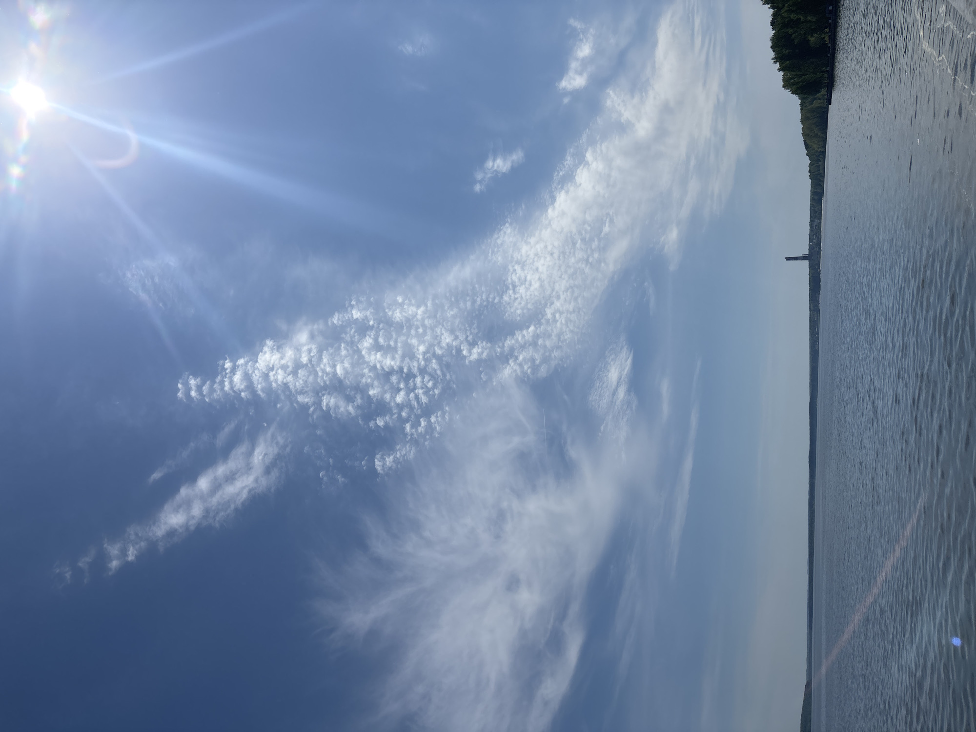 The image shows a daytime view of the Kama River in Chaikovsky, Russia. The sky is predominantly blue with various types of clouds, including cirrus and cirrocumulus. The sun is visible in the upper left corner, creating a bright glare. The river fills the right side of the frame, with a dark shoreline visible in the background, suggesting the opposite bank. A tall structure, potentially a tower or smokestack, is barely visible on the distant shoreline.
