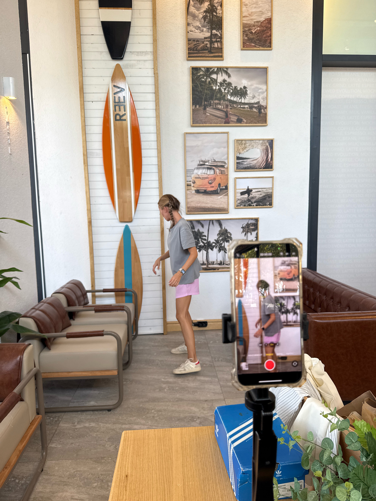 In Antalya, Turkey, a young woman with braided hair, wearing a grey t-shirt and pink shorts, is standing in a cafe with a modern, beach-themed decor. She is in the process of filming a video on her smartphone, which is mounted on a tripod. The cafe's walls are adorned with framed pictures of beaches, palm trees, and surfboards, with two large decorative surfboards also mounted on the wall behind her. Brown leather and beige upholstered seating is arranged around a light-colored wooden table in the foreground.