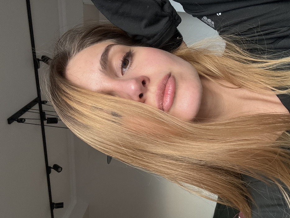 A close-up, chest-up image depicts a young adult female indoors. She has long, straight, light brown hair, parted to her right, with a portion covering the left side of her face, partially obscuring her left eye. Her right eye is visible, with dark lashes and defined eyebrows. Her lips are full and closed. She wears a black long-sleeved garment, possibly athletic wear, exhibiting a subtle hexagonal pattern on the right arm/shoulder area. Her left hand is raised, positioned to the left and slightly behind her head, causing a minor pull on her garment. The background shows a light-colored ceiling with a black track lighting system featuring multiple spotlights. The precise location within the city of Unknown, Unknown, is not discernible from the image.