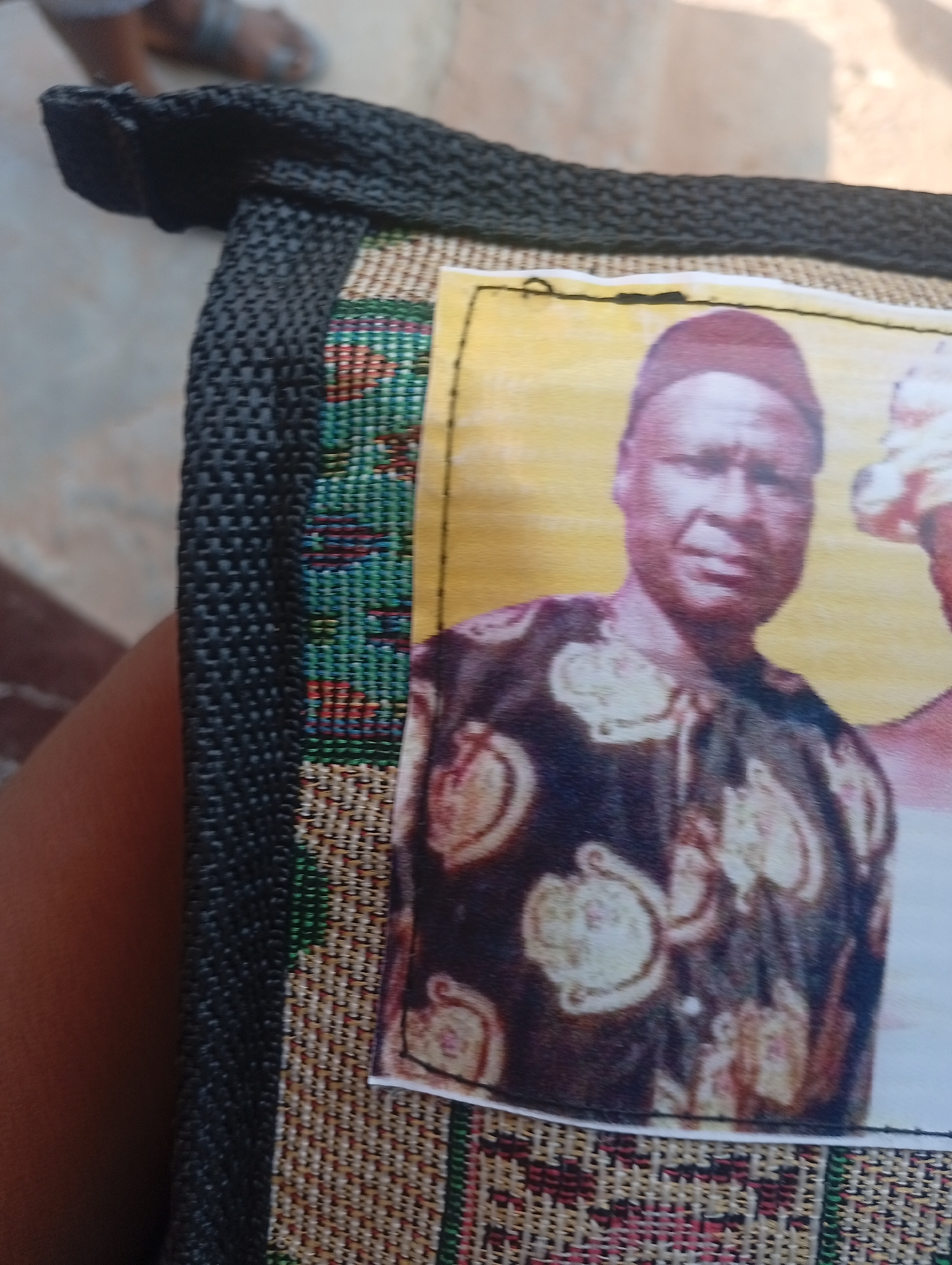 The image shows a close-up of a dark woven fabric bag with a patterned fabric inset. A photograph is affixed to the patterned inset. The photograph shows a man wearing a dark, patterned tunic and a cap. He is looking directly at the camera. To his right, partially visible, is another person wearing a white headwrap. The overall color palette of the photograph is warm, with a yellow background. The setting appears to be outdoors, suggested by the lighting. The fabric of the bag is rough and textured, with a black woven exterior and a colorful, intricate woven pattern beneath the photograph. The background is blurred, but appears to be a paved surface.