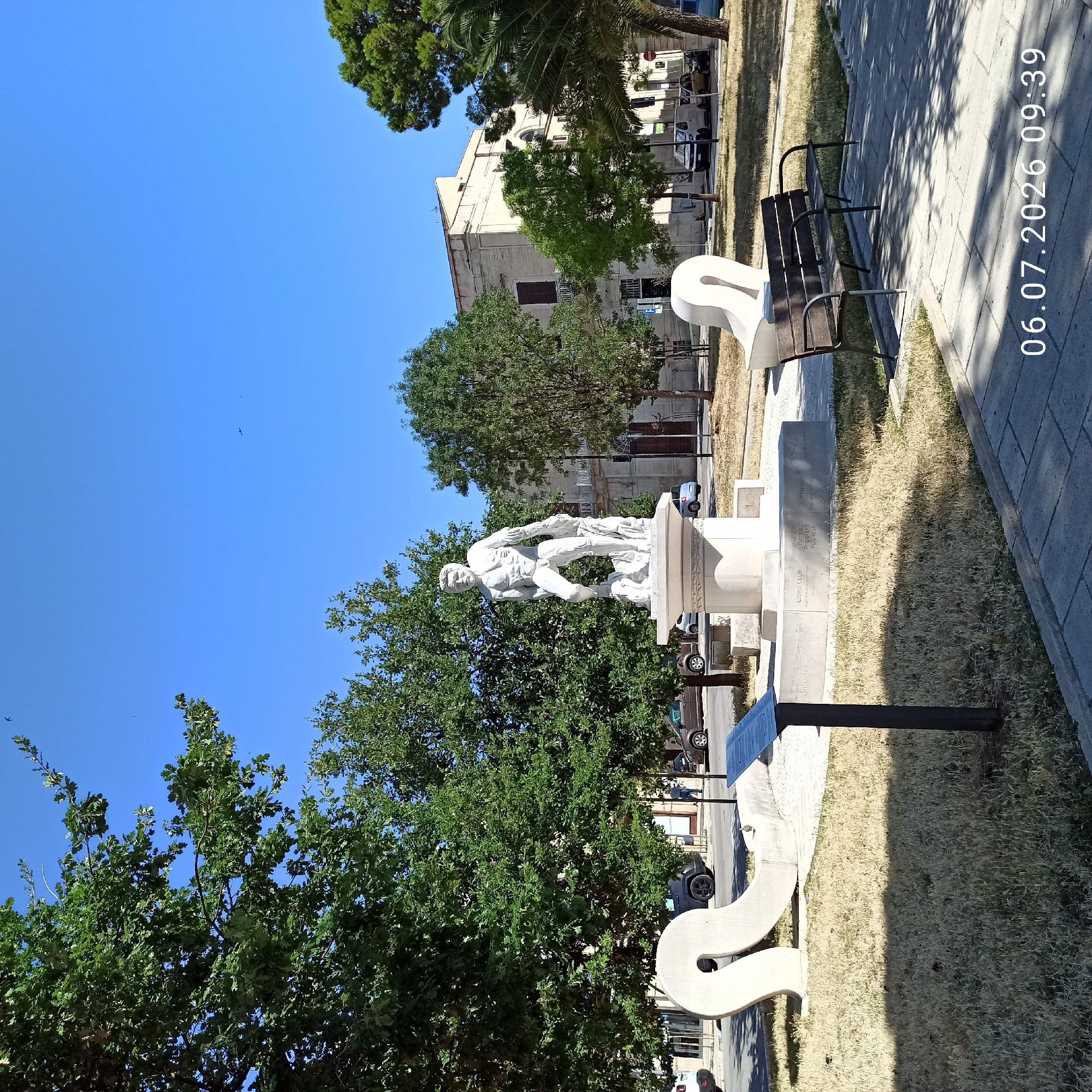 The image depicts a sunlit public square in Ruvo di Puglia, Italy, featuring a white marble statue of a muscular, nude male figure standing on a pedestal. The statue is positioned in the center of the frame, surrounded by green trees and dry grass. In the foreground, there are two abstract white sculptures and a dark wooden park bench. A metal informational plaque is also visible near the dry grass. In the background, a light-colored building with classical architectural elements and a balcony can be seen, along with trees and parked cars on a street. The sky is clear and blue, with a few small birds visible. The timestamp "06.07.2026 09:39" is overlaid in the lower right corner.