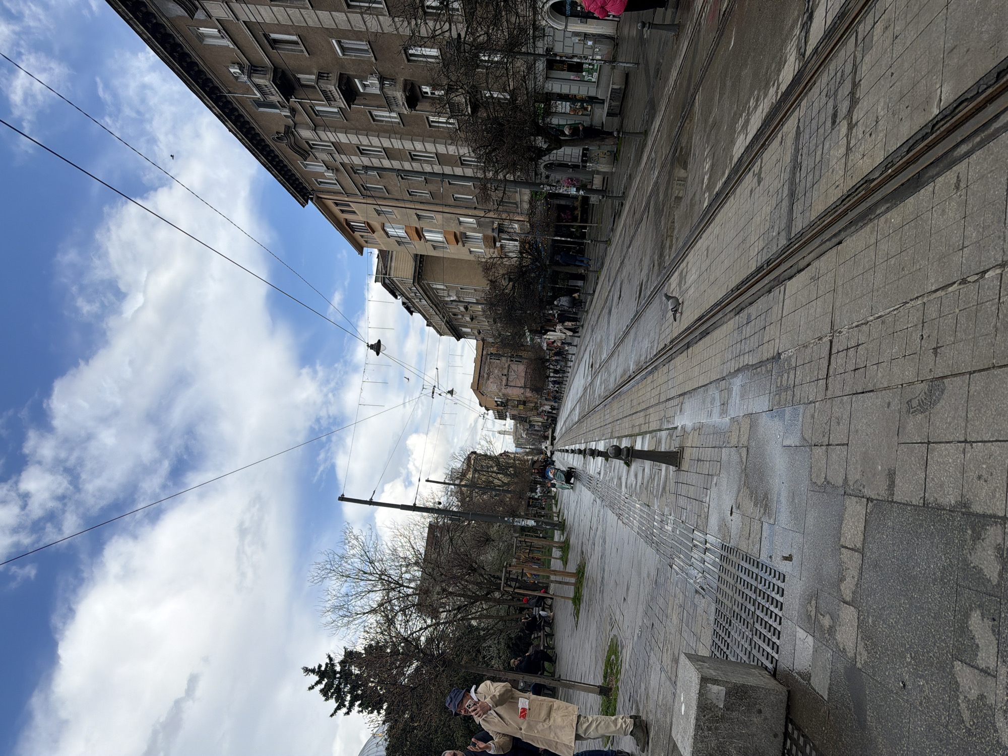 The image captures a street scene in Sofia, Bulgaria, characterized by tram tracks running along the pedestrian walkway. Buildings line one side of the street, displaying a mix of architectural styles. Bare trees interspersed with benches are visible on the sidewalk, and several people are seen walking and sitting. Overhead wires span across the street, likely powering the trams. The sky is partly cloudy. The pavement appears wet, and the overall lighting suggests it is daytime.