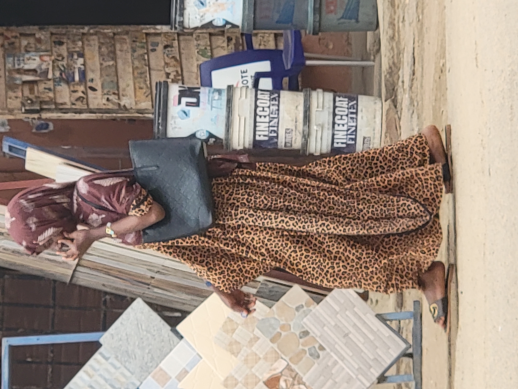 This media file captures an outdoor scene, likely a market or commercial area, in bright daylight in Maiburiji, Nigeria. The central subject is a person, likely a woman, wearing a long, loose leopard-print dress and a maroon head covering. She is carrying a dark-colored shoulder bag and has bare feet in sandals. Her left hand is raised towards her face, possibly holding a phone or touching her cheek.
Behind the person, several white buckets are stacked, some with blue lids. Visible text on these buckets includes "FINECOAT" and "FINETEX", indicating they might be paint or building material containers. A blue plastic object is also partially visible behind the buckets, with the partial word "OTE" (likely from "VOTE") visible on it.
To the left of the person, various patterned floor tiles are displayed on a rack, suggesting a building materials vendor or shop. The background features a weathered structure, possibly a wall made of wooden planks or corrugated metal, covered in faded papers or posters. The ground appears dusty, consistent with dry, outdoor conditions. The overall setting suggests a bustling, everyday street or market scene.