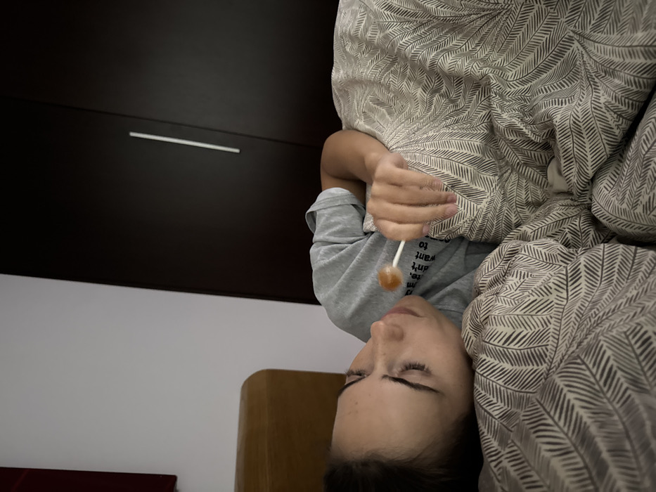 An individual is depicted lying on their left side in a bed, indoors. The person, appearing to be a young adult, has dark hair and fair skin with visible freckles, and is wearing a light grey short-sleeved t-shirt with partial dark text on the chest. The individual holds an amber-colored, spherical lollipop with a white stick in their right hand, positioned near the mouth. The head and upper torso are supported by a patterned pillow, matching the duvet covering the body, both featuring dark, stylized leaf or feather motifs on a light background. Behind the individual's head, a light brown wooden headboard with a rounded top edge is visible. The background consists of a white wall on the upper left and a dark brown wardrobe with a single vertical silver handle occupying the right side of the frame. A small red object is partially visible in the upper left corner of the scene. The image contains no discernible landmarks or identifiable features to confirm the specific location as Warsaw, Poland.