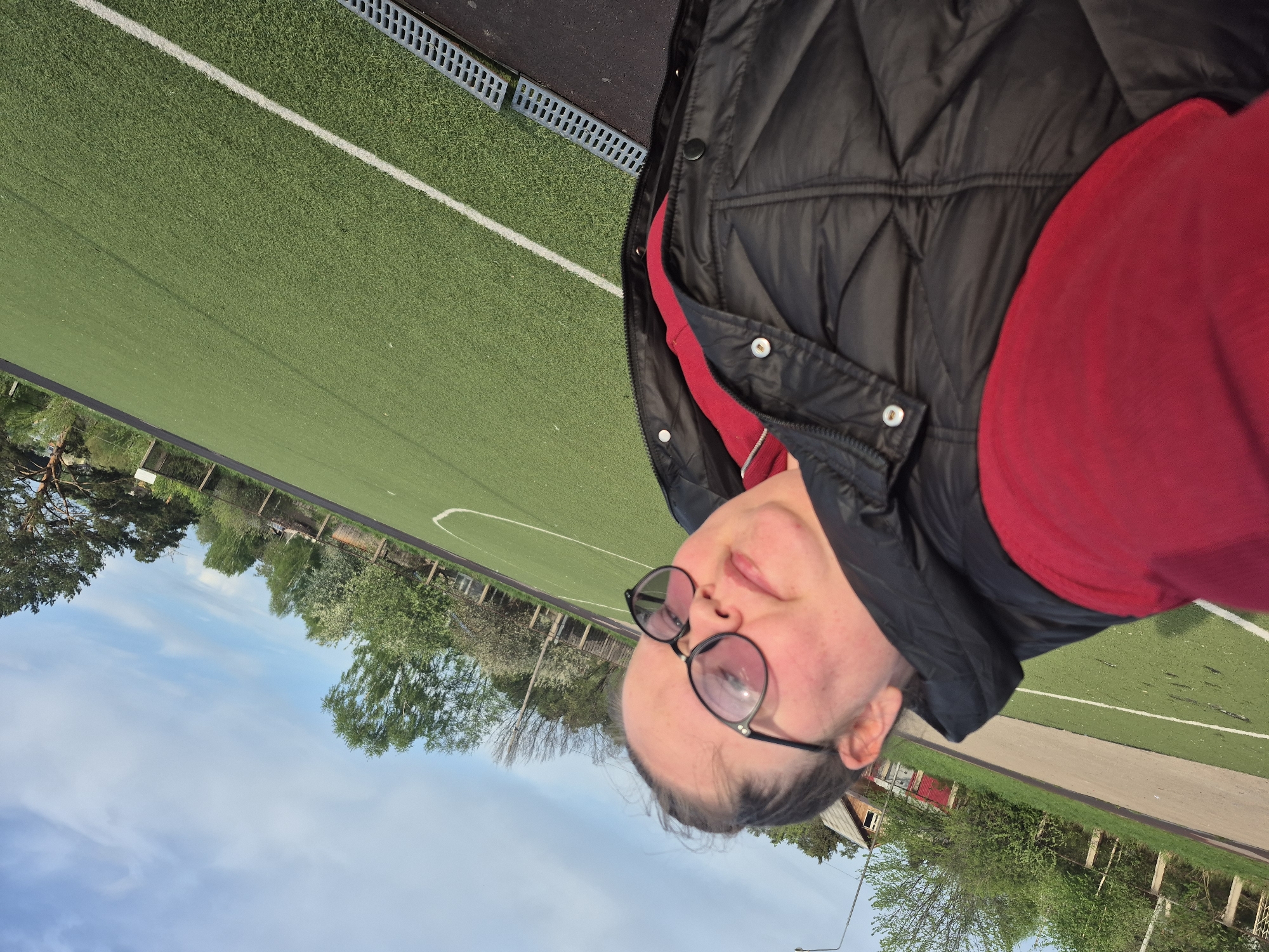 The image shows a person wearing glasses, a red shirt, and a black vest, possibly in the city of Unknown, Unknown. The background includes a green artificial turf field with white lines, trees, a blue sky with clouds, and buildings.