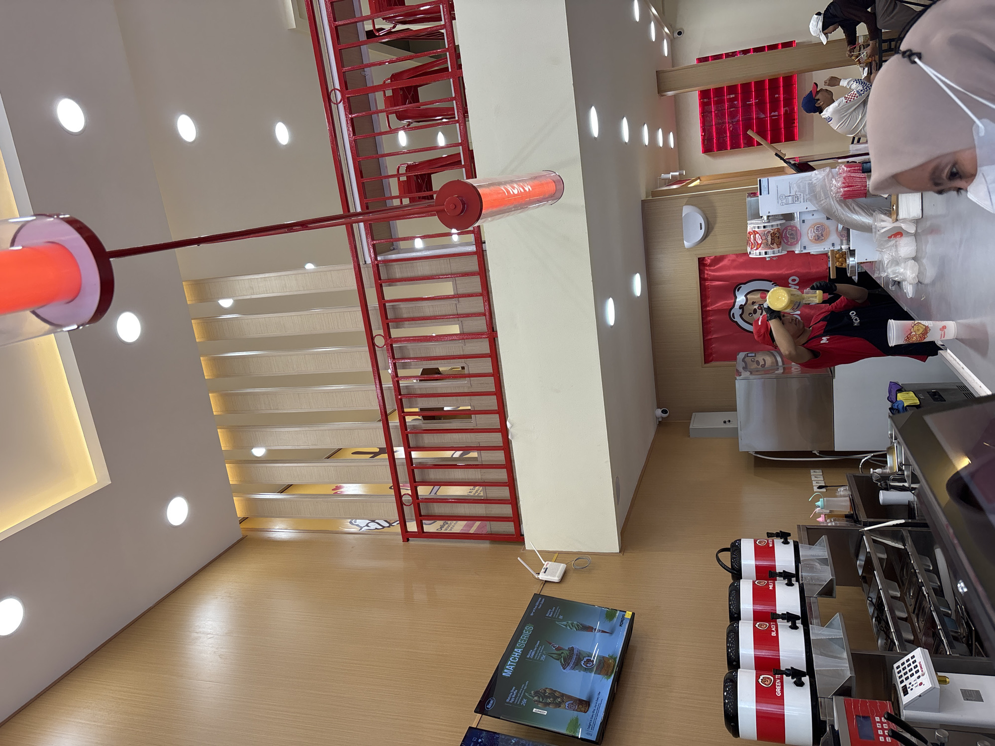 The image captures the interior of a Honu store in North Jakarta, Indonesia, focusing on its beverage preparation area. A person wearing a red uniform is actively preparing a drink. A customer, identifiable by their hijab, is seen in the foreground. In the background, other customers and store personnel are visible. The store's design incorporates red metal framework, including a ladder, and a sign advertising "Matcha Series." The beverage preparation area includes beverage dispensers, a counter, and other equipment.