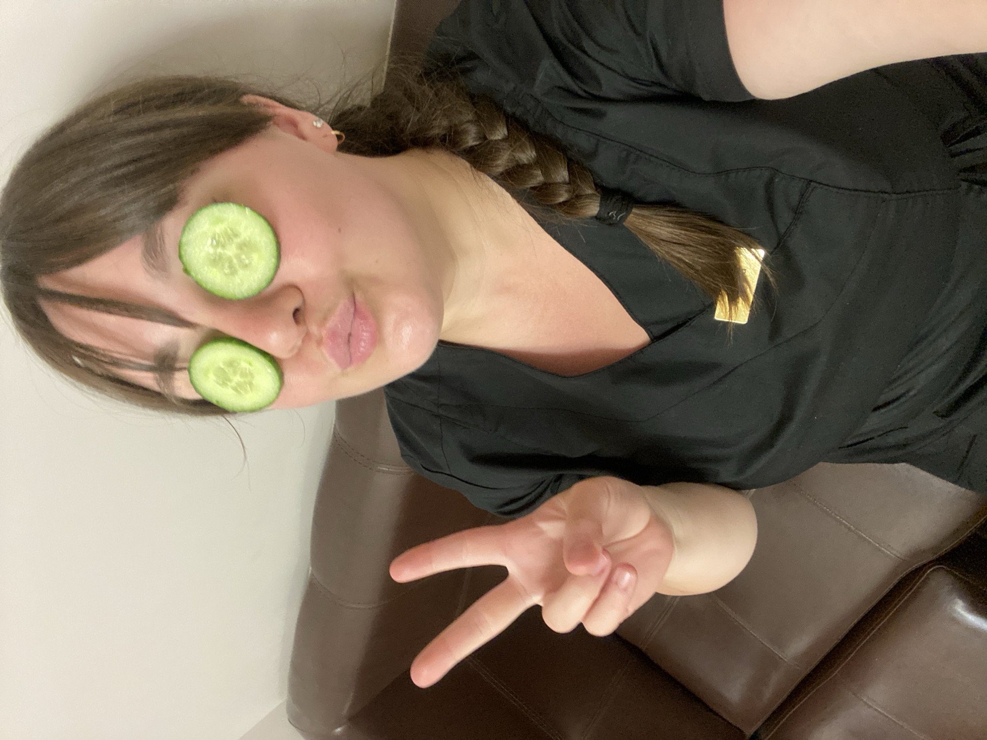A woman is pictured, with a braid extending to her left. She has cucumber slices over her eyes and makes a peace sign with her right hand. She is wearing a dark colored top, and is sitting on a leather couch. The location is not discernible from the image and is therefore unknown.
