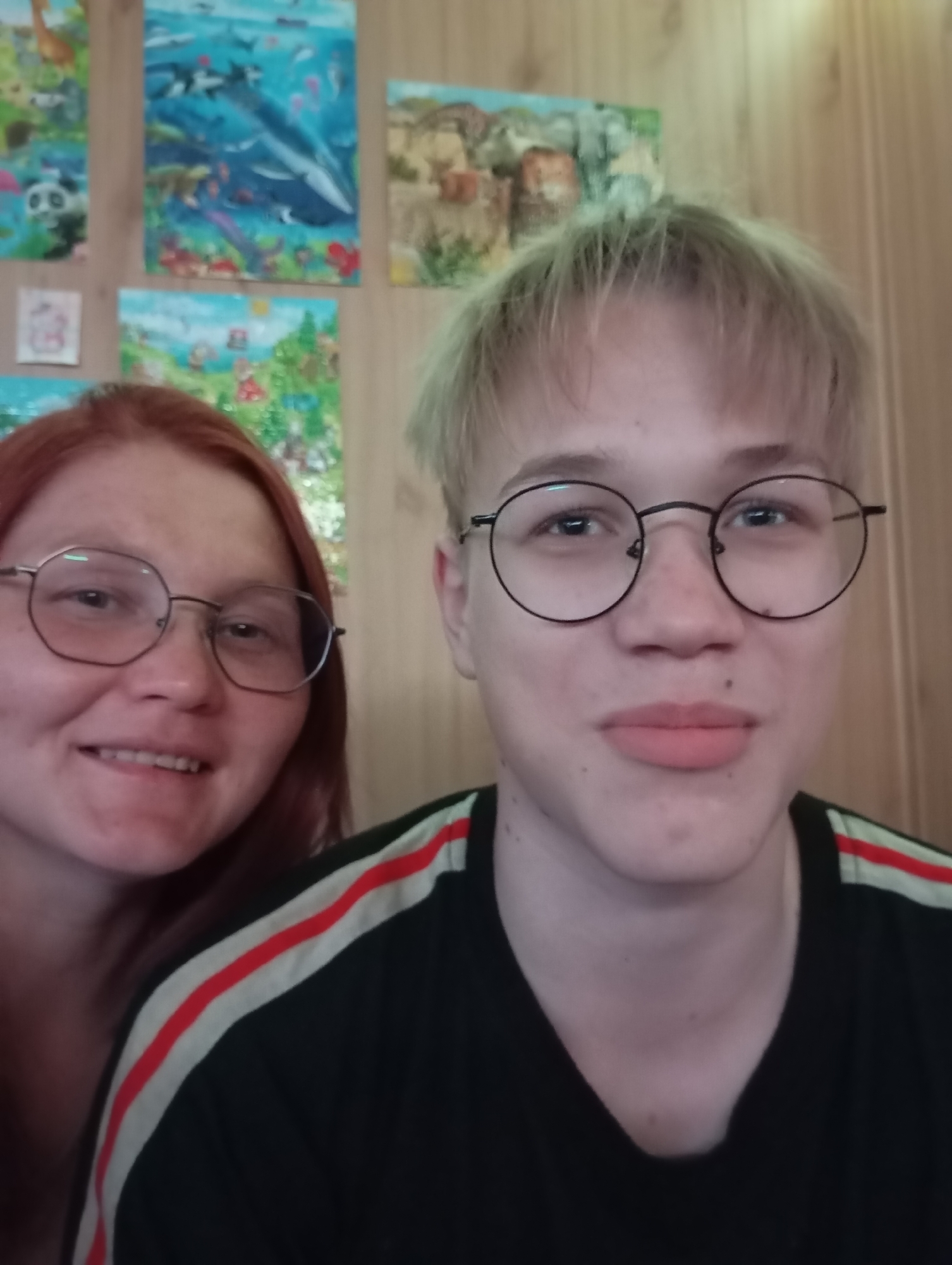 The image shows two people in an indoor setting. The person on the left has red hair and is wearing glasses. They are smiling slightly. The person on the right has light-colored hair, is also wearing glasses, and has a neutral expression. The person on the right is wearing a black t-shirt with red and white stripes on the shoulder. Behind them are several children's paintings on the wall. The location is Unknown, Unknown.